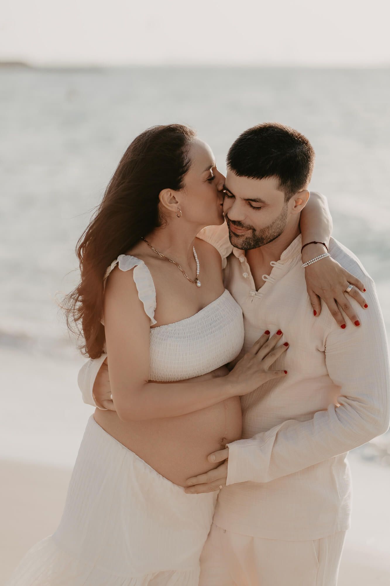 A romantic couple in white embraces on the beach during a soft sunset photoshoot.