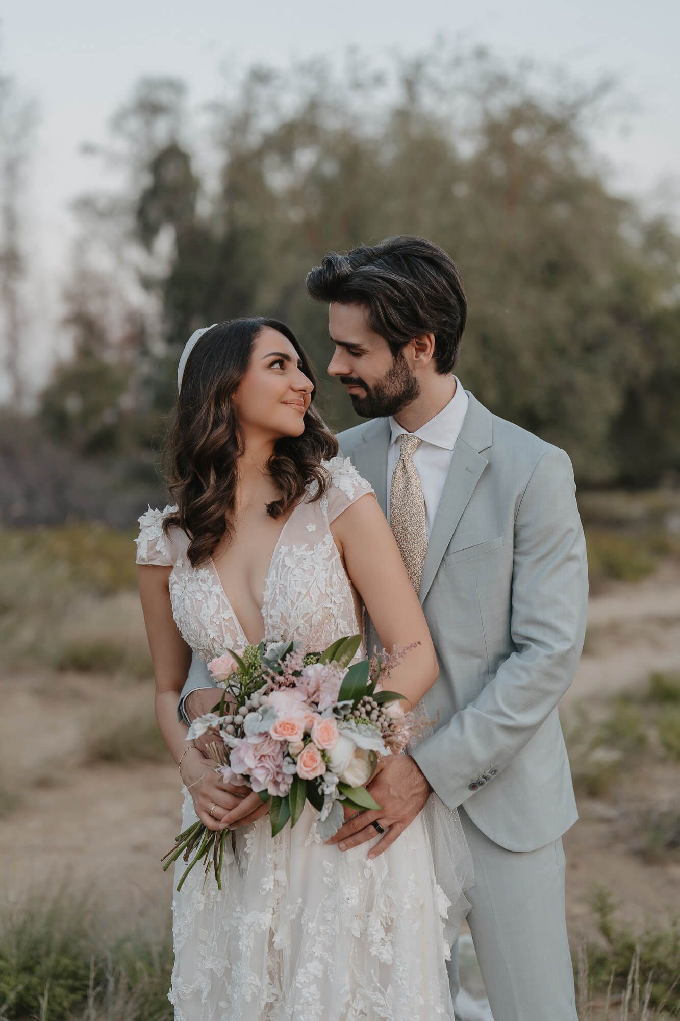 Couple in wedding attire embrace outdoors while holding a soft pink and white floral bouquet against a natural background.