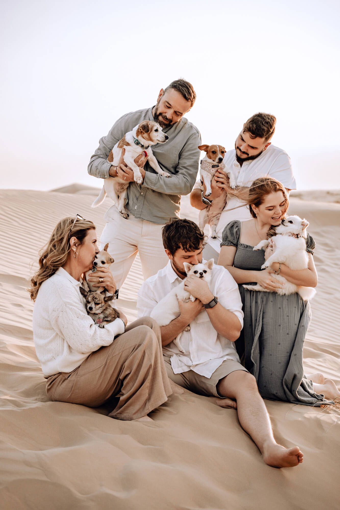 Casual family portrait with multiple small dogs sitting together on sand dunes during sunset.