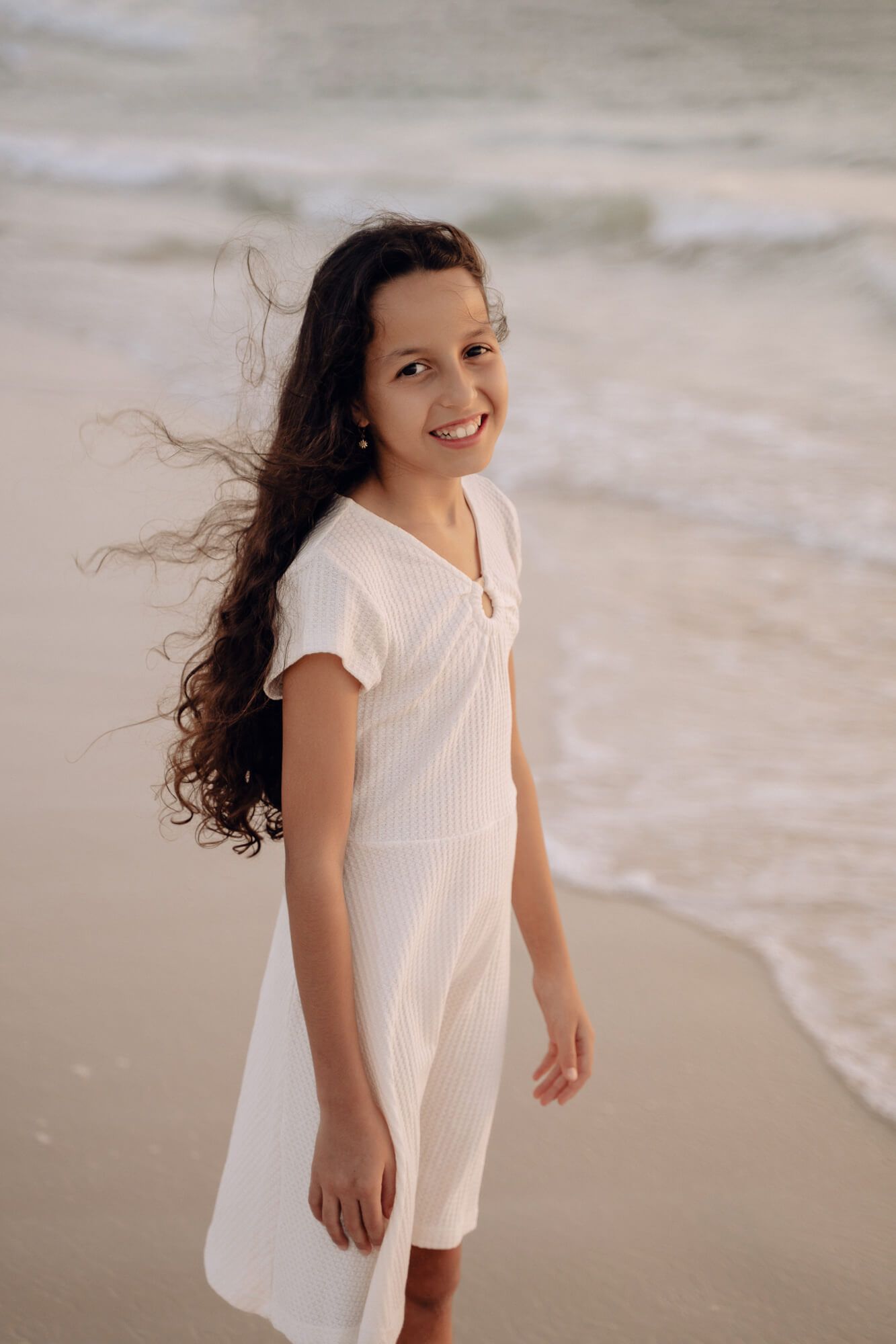 A person with flowing hair smiles brightly while standing on a sandy beach at sunset.