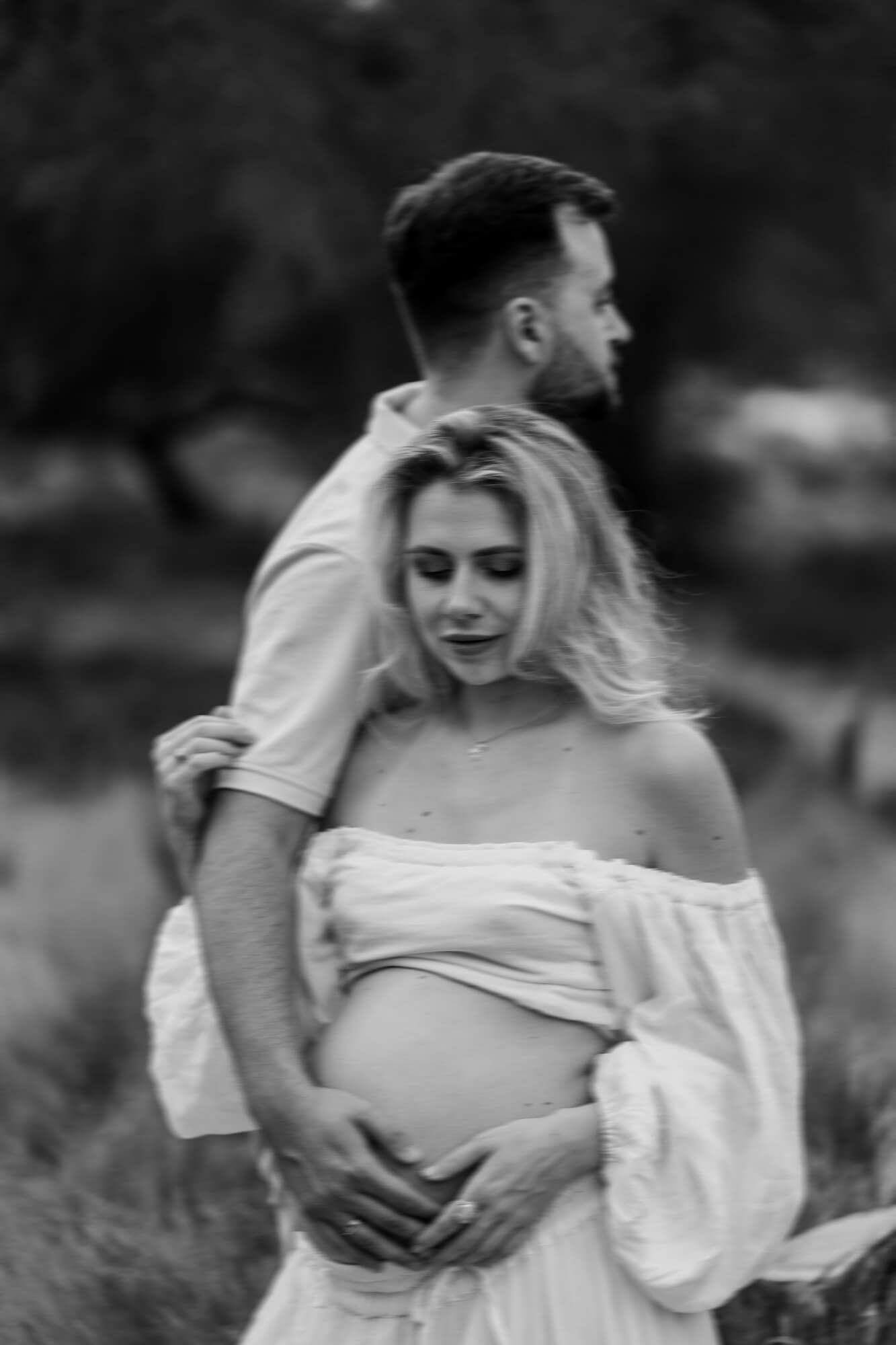 Black and white maternity photo captures intimate moment between couple in outdoor setting.
