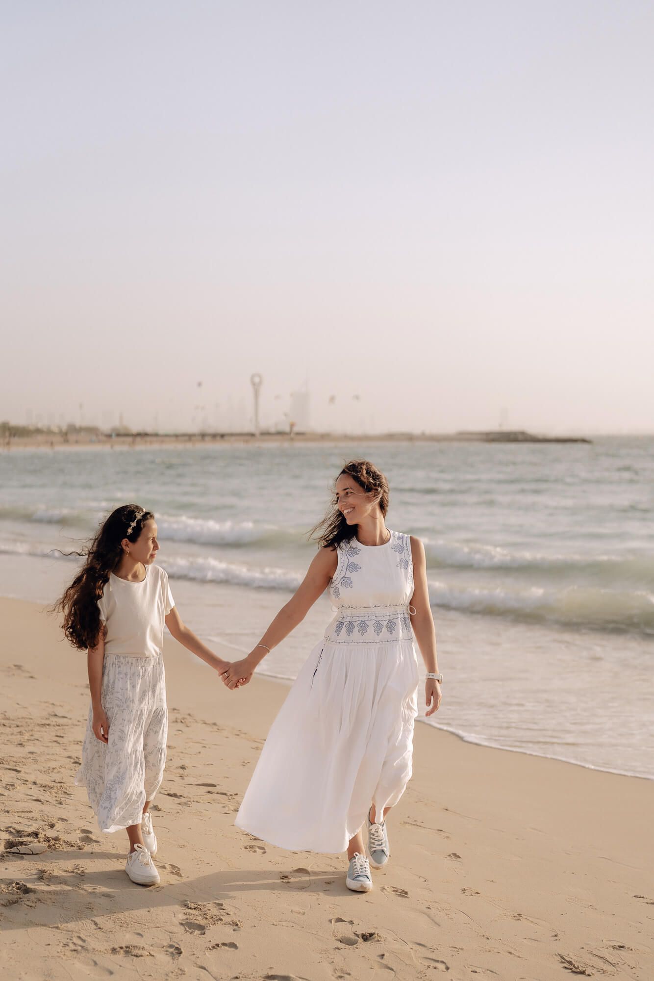 Two people in flowing white dresses walk hand in hand along a beach with a cityscape in the background.
