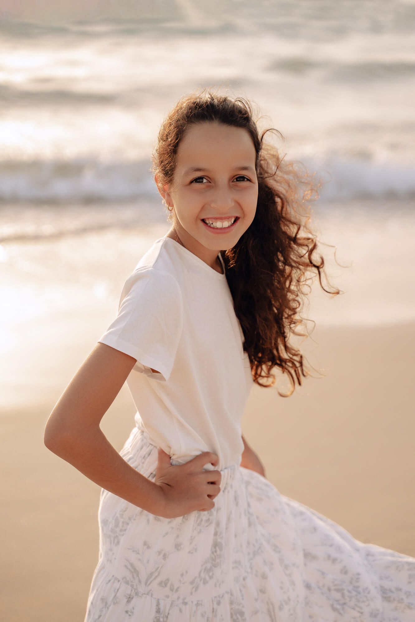Young girl with curly hair laughing and playing in a white dress on the beach.