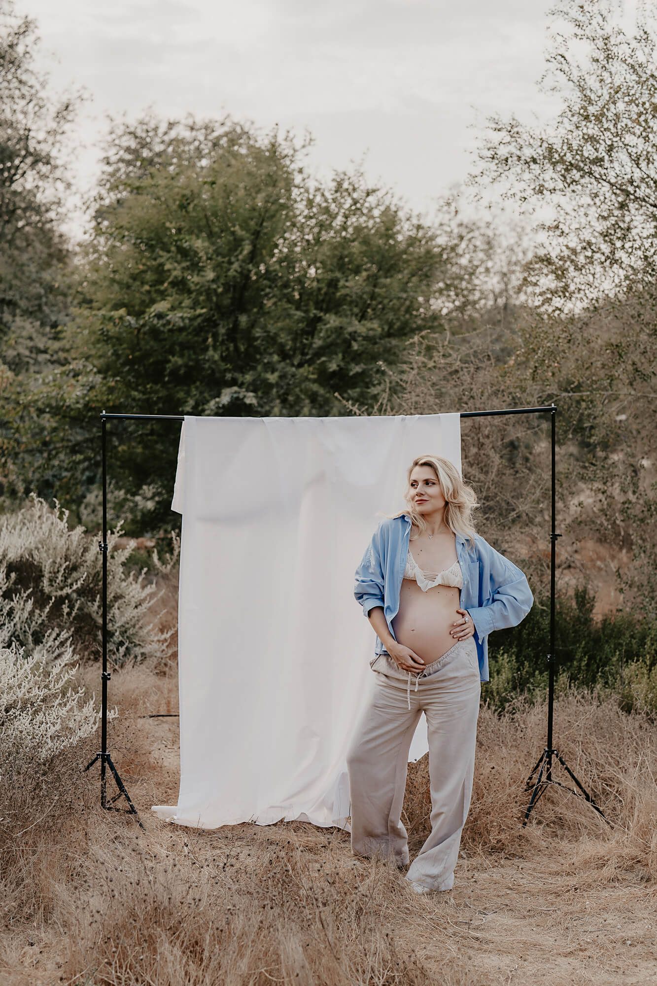 Outdoor maternity portrait with white backdrop showing a relaxed pose in denim and beige attire.