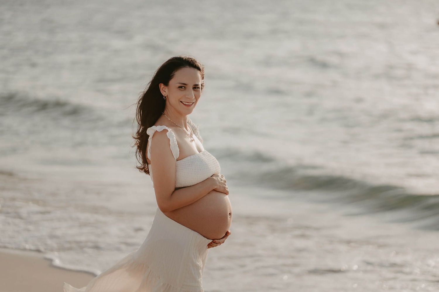A pregnant woman in a white dress stands on a sandy beach at sunset, smiling while cradling her baby bump.
