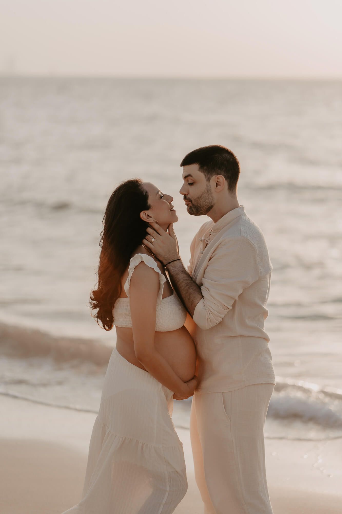 A romantic couple in white outfits share an intimate moment on the beach at sunset.