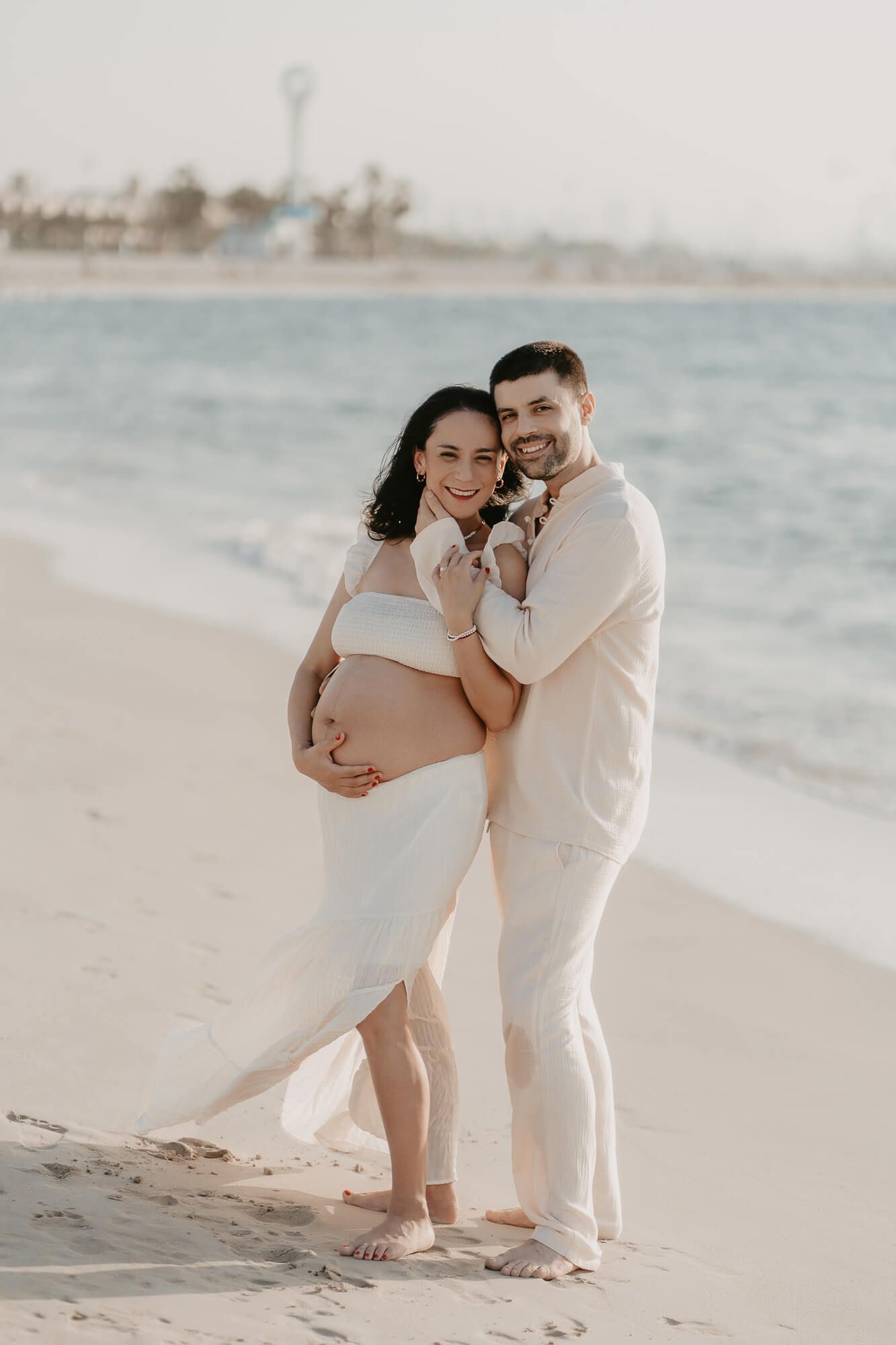 A romantic couple in light colored clothing embraces and dances together on a sunny beach.