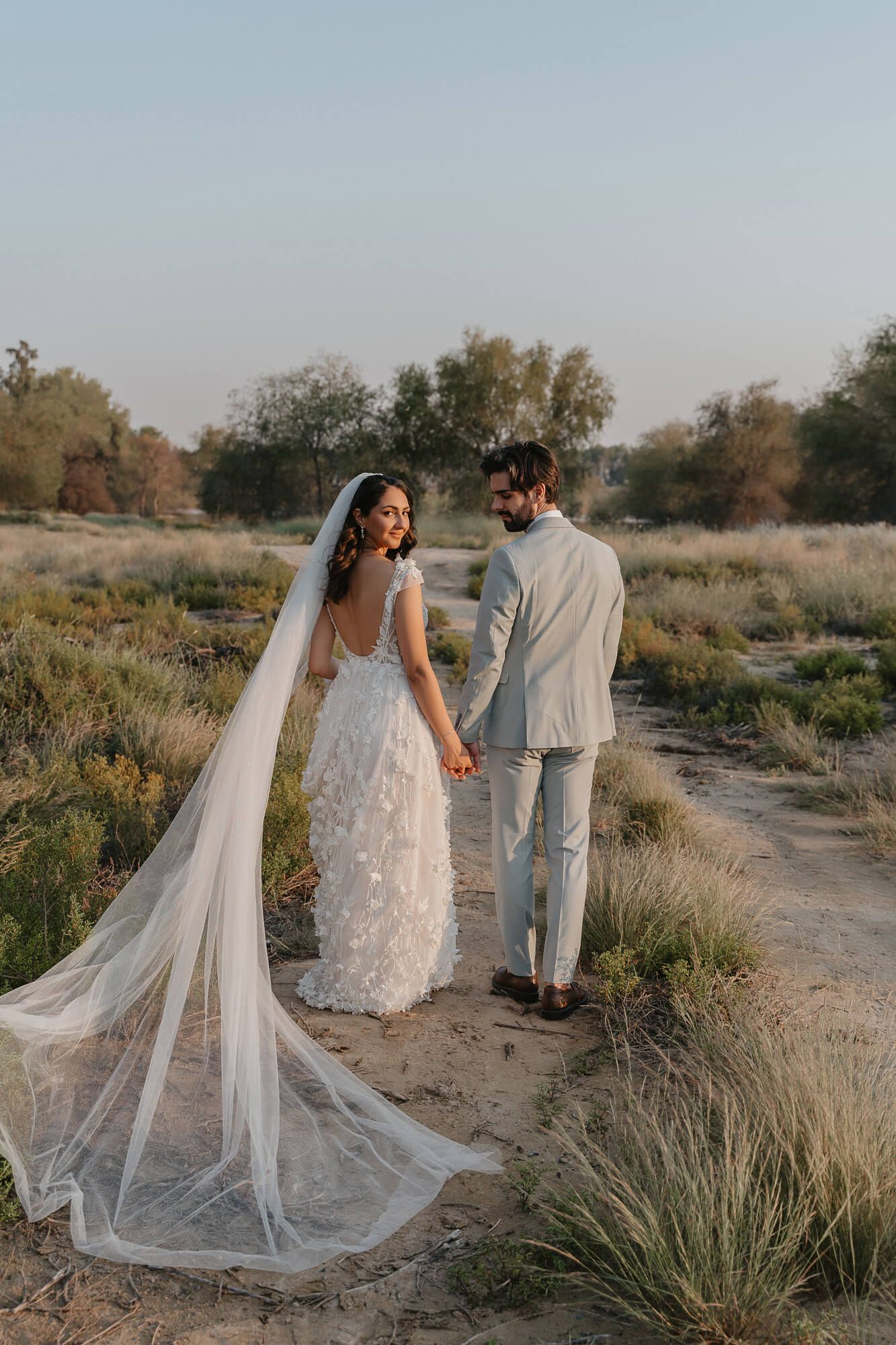 A couple in wedding attire stand together on a dirt path during golden hour, with a long flowing veil trailing behind.