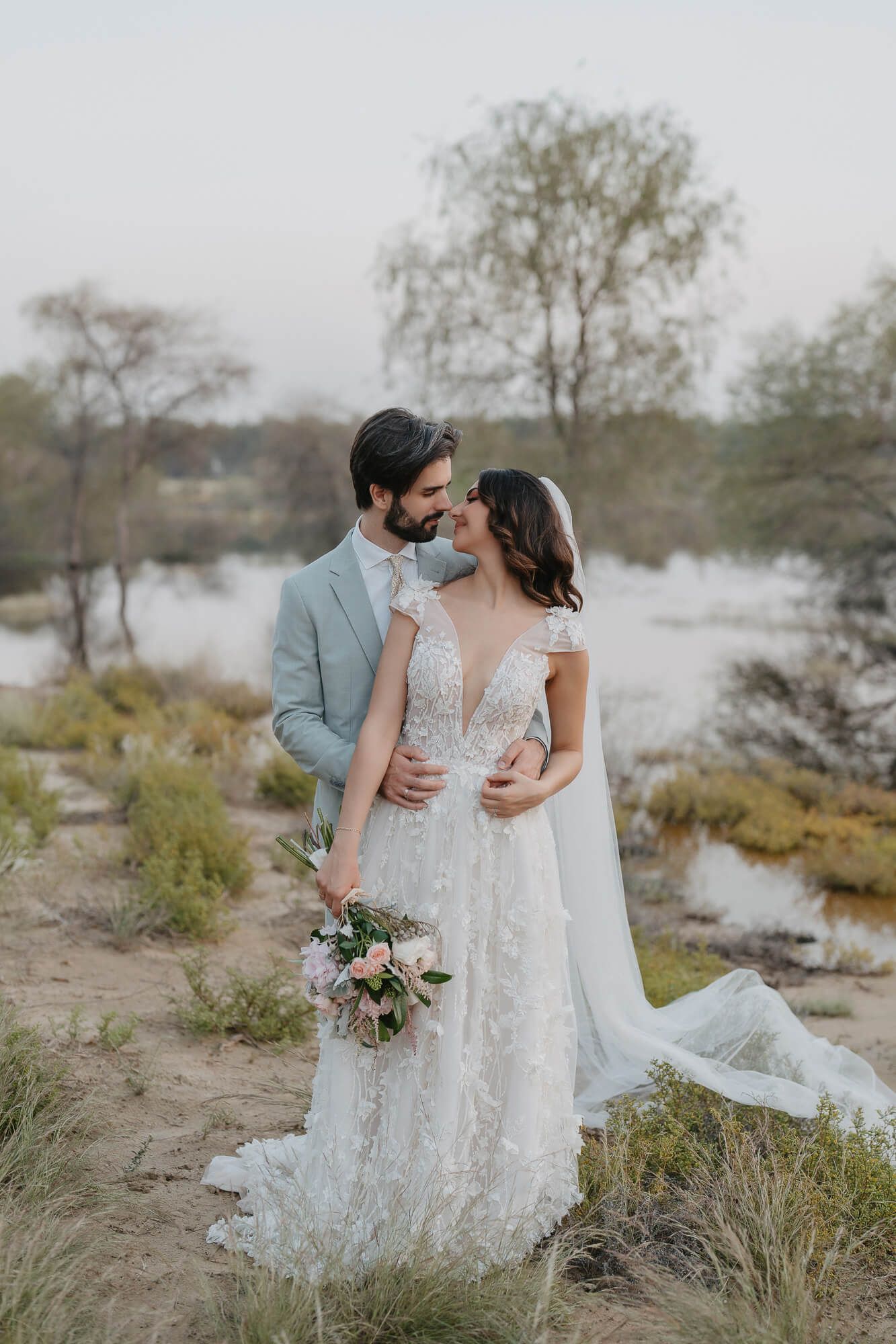 A couple in wedding attire embraces on a misty beach with coastal vegetation and soft neutral tones in the background.