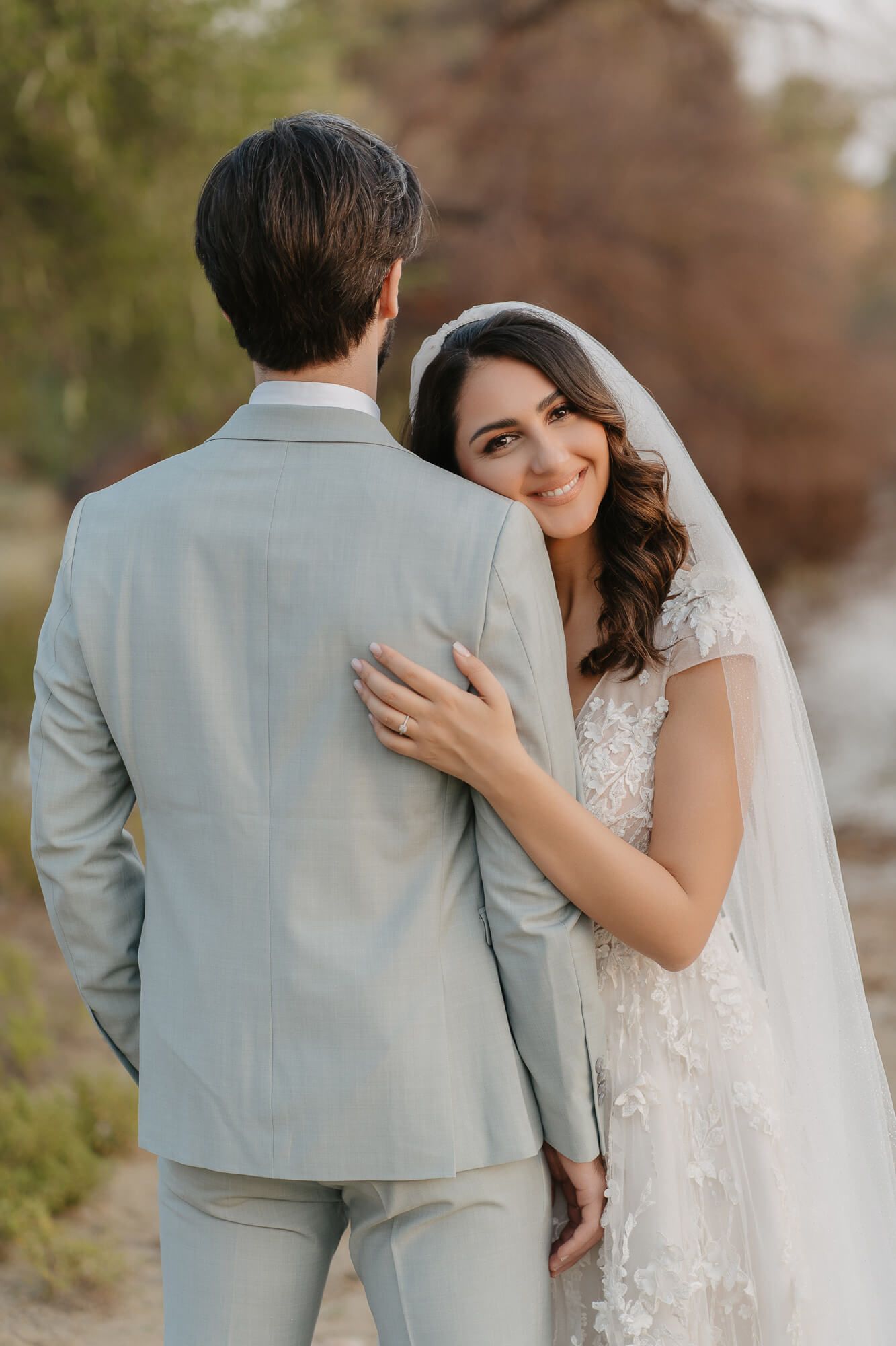 Bride in white lace dress and veil embraces groom in light gray suit during outdoor wedding photo session.