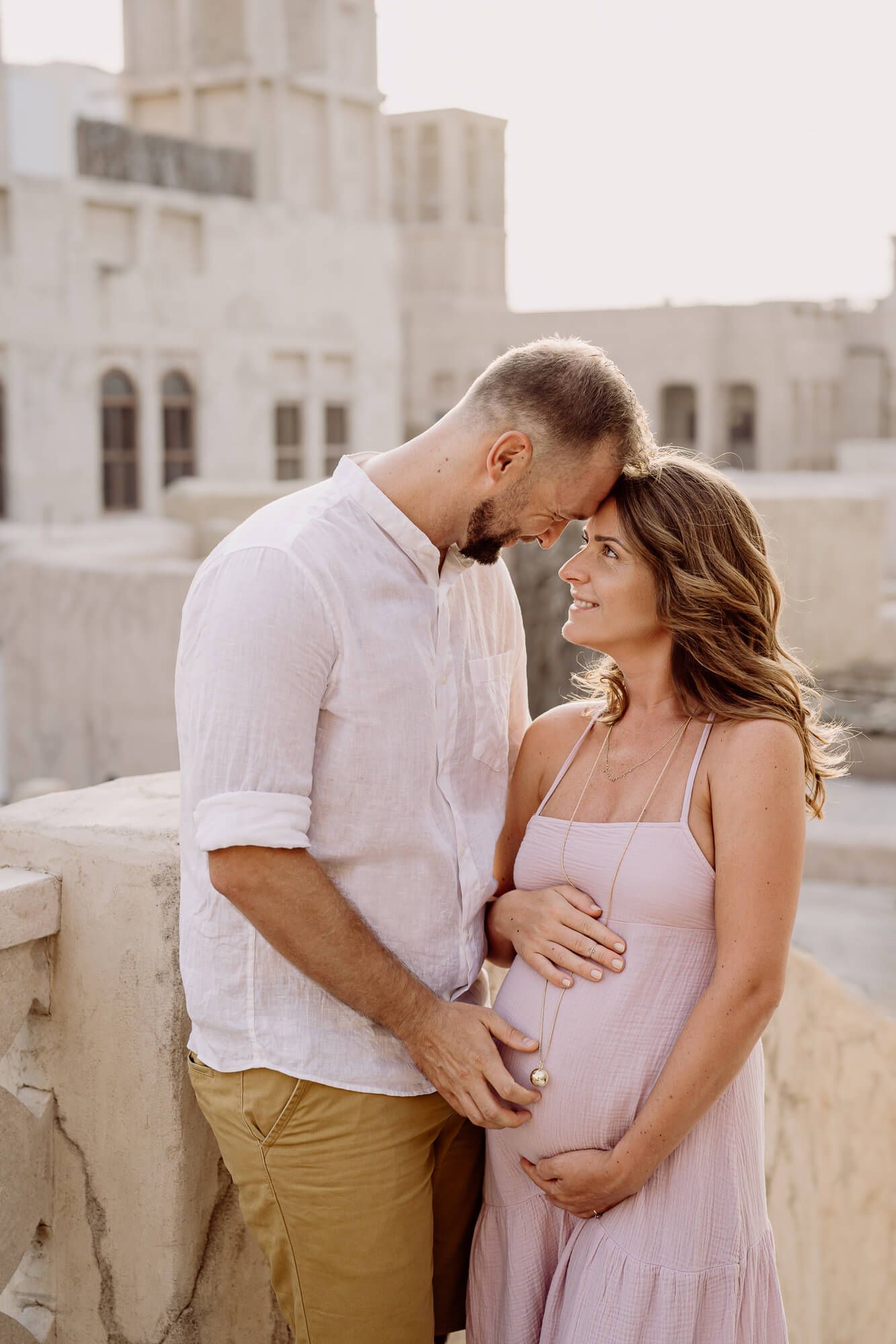 A romantic couple embraces on a rooftop terrace, the woman wearing a pink dress and cradling her pregnant belly.