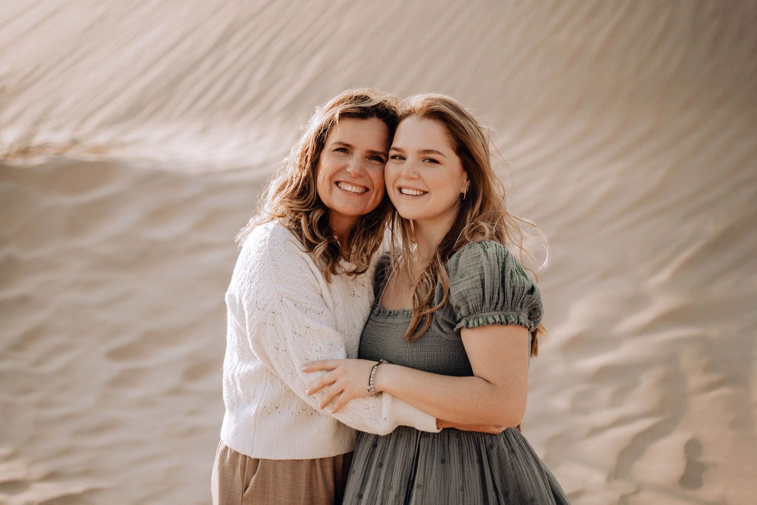 Two people smile and embrace while posing for a portrait against sandy desert backdrop.