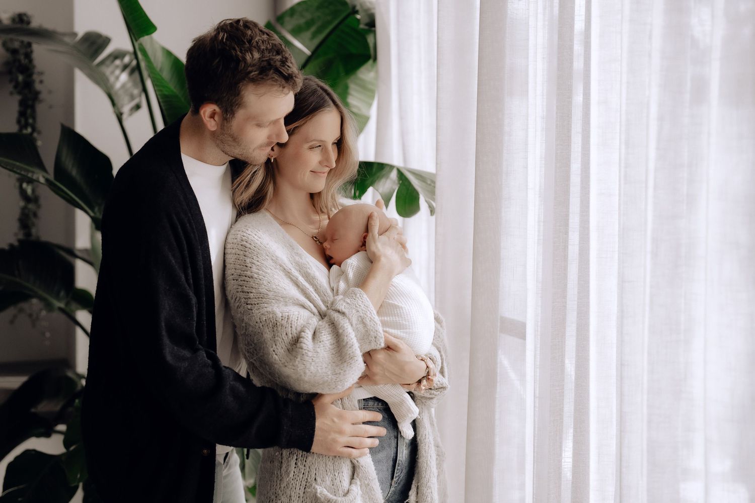 Intimate indoor portrait of an expecting couple embracing by a window with greenery.