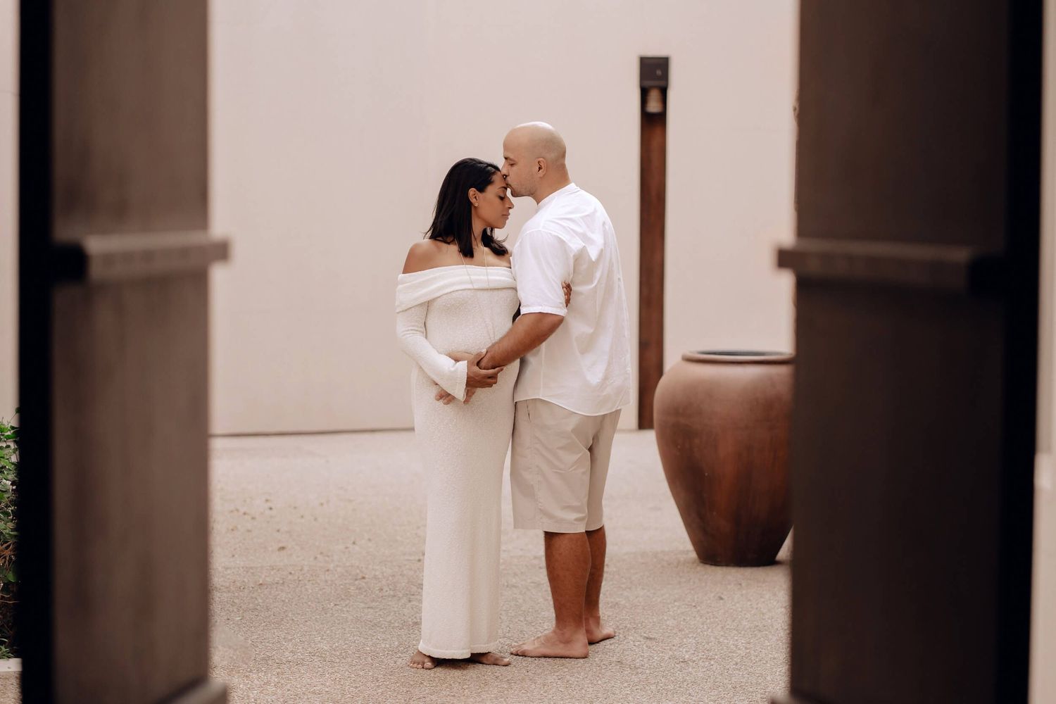 An intimate maternity photo of a couple in white clothing embracing in a covered walkway.