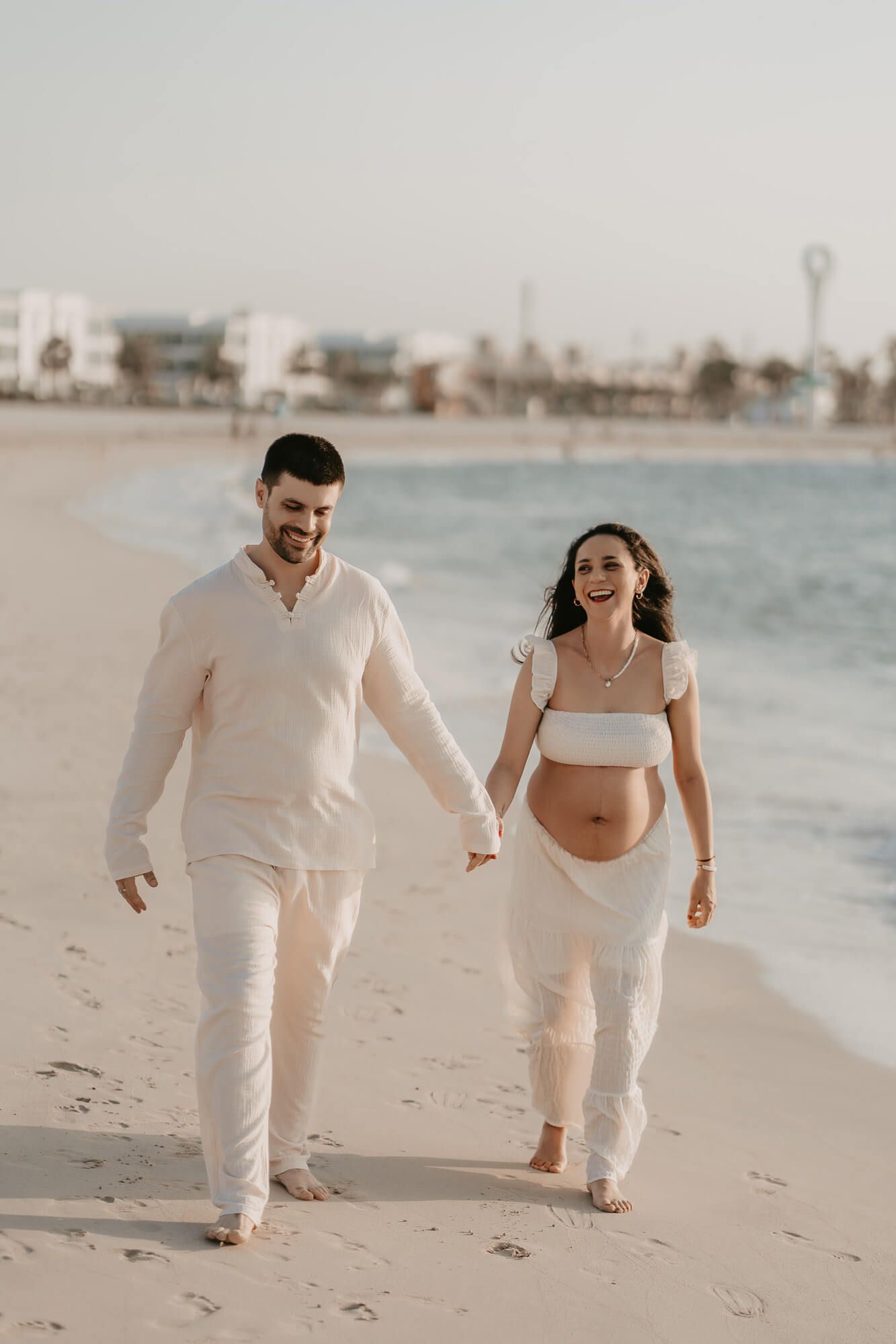 A couple walks hand in hand on a beach at sunset, wearing all white outfits with the woman showing her pregnant belly.