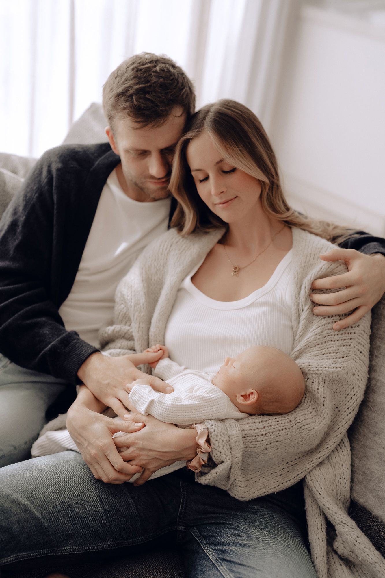 Intimate family moment with parents tenderly holding their sleeping newborn baby on a couch near window light.