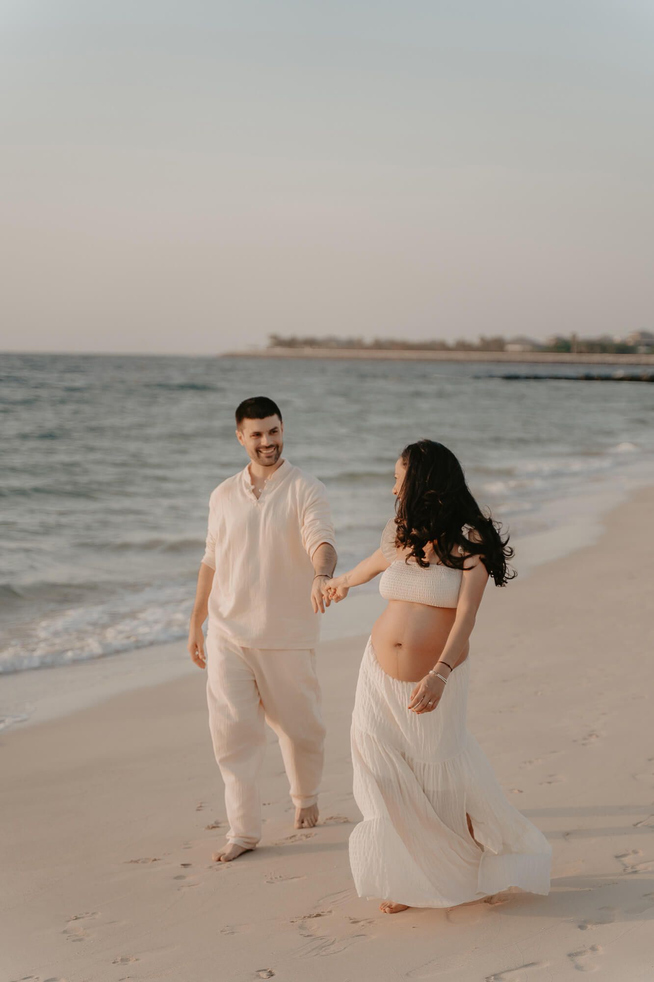 Couple in white outfits walking and dancing playfully together on a sandy beach at sunset by the ocean.