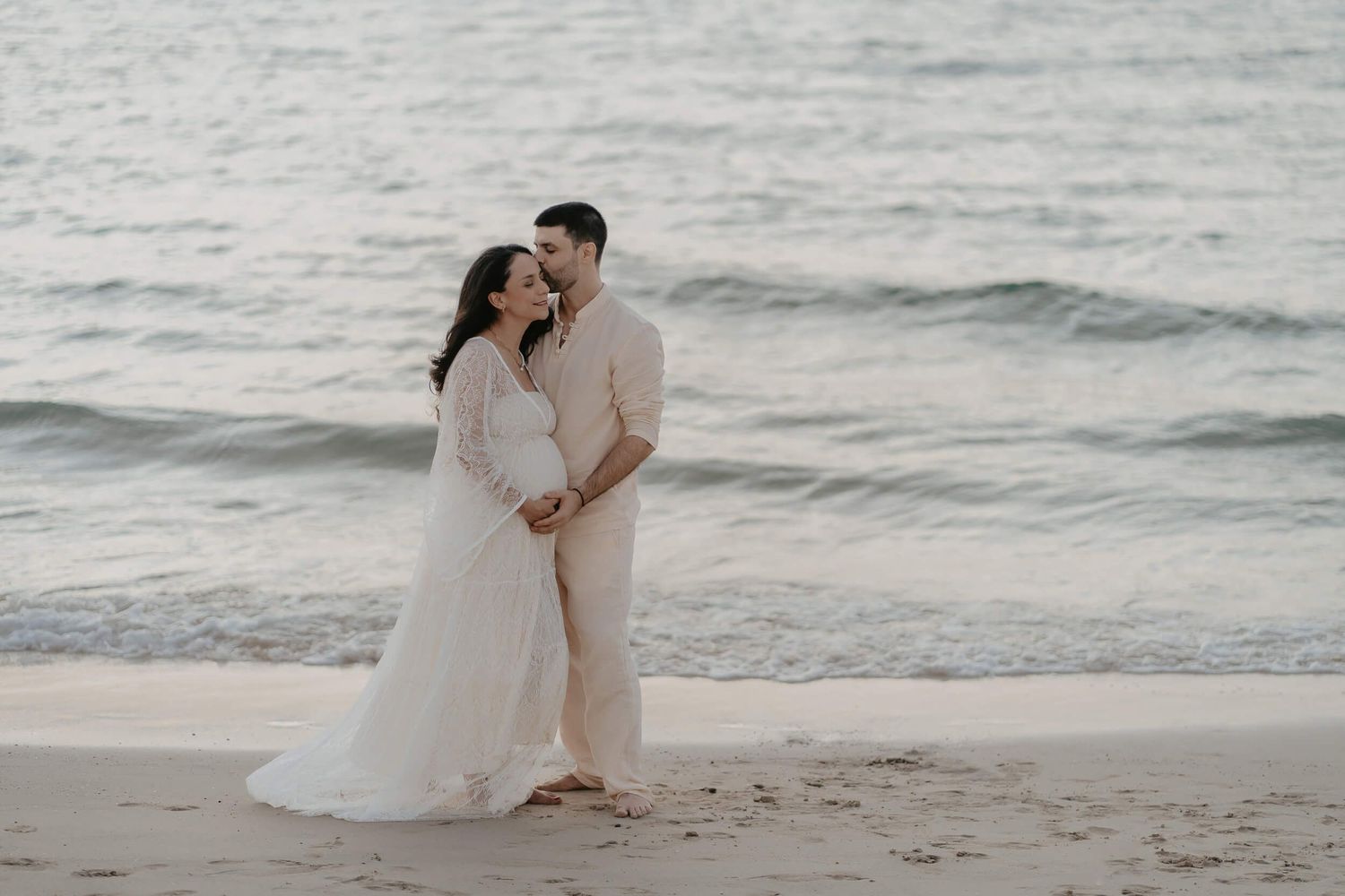 A romantic beach wedding photoshoot with a couple embracing as gentle waves roll onto the shore at dusk.
