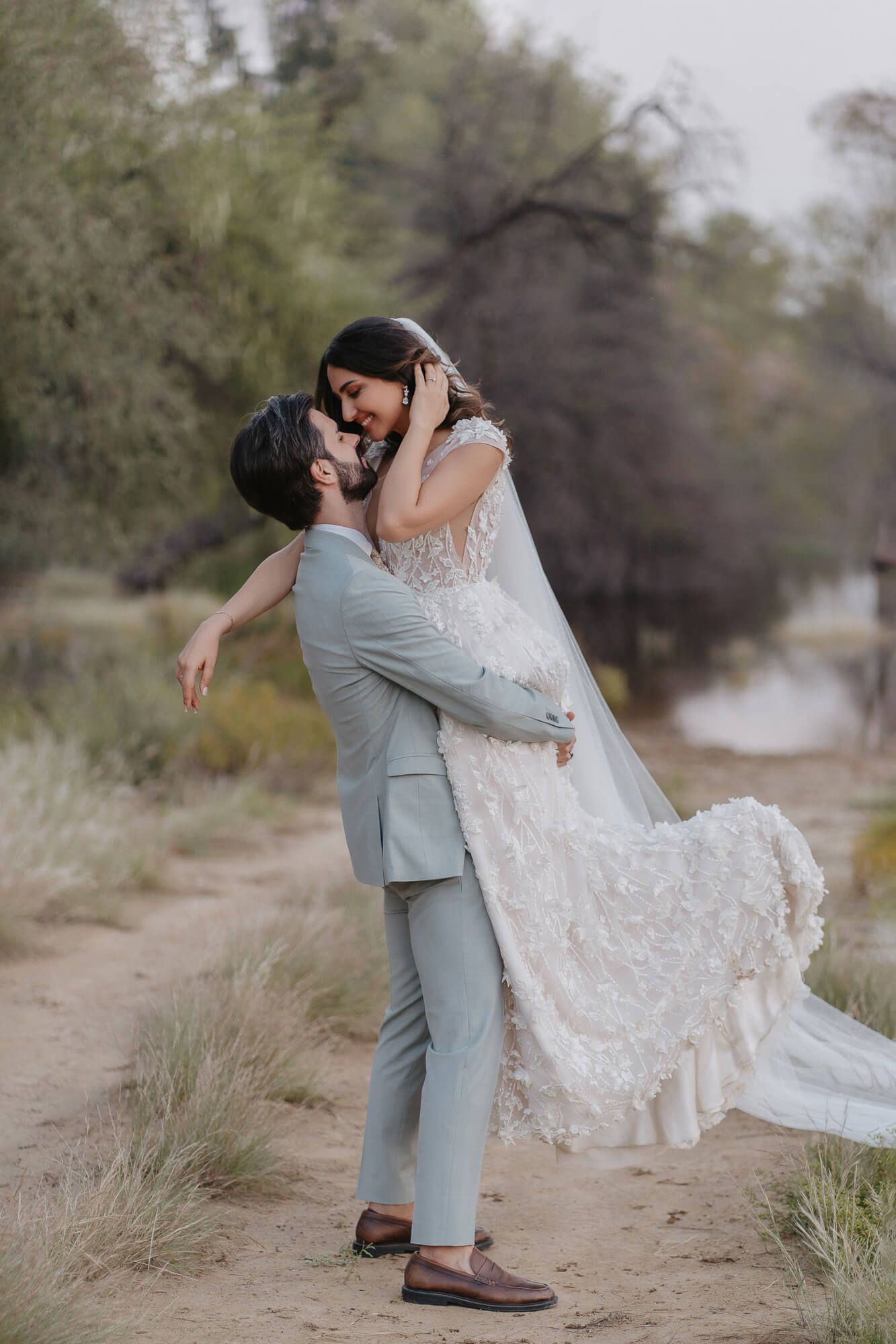 A couple embraces and dances together on a sandy desert path surrounded by desert brush.