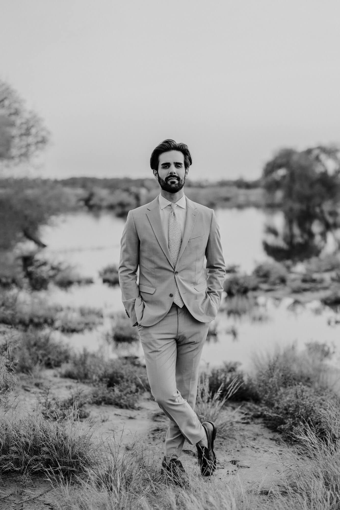 Stylish groom in light suit walks through desert brush in black and white photography session.