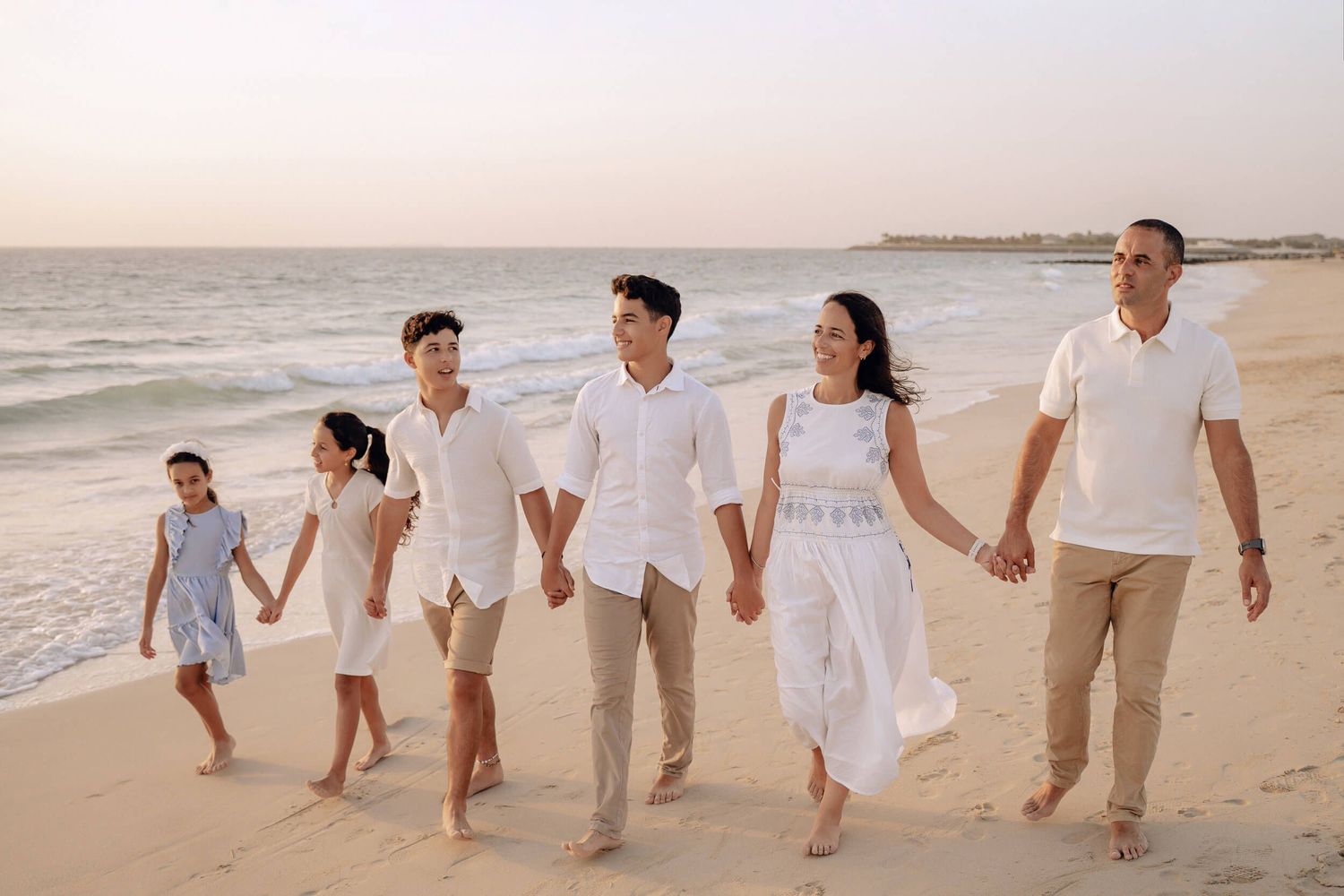 A group wearing white and beige clothing walks together joyfully along a sandy beach at sunset.