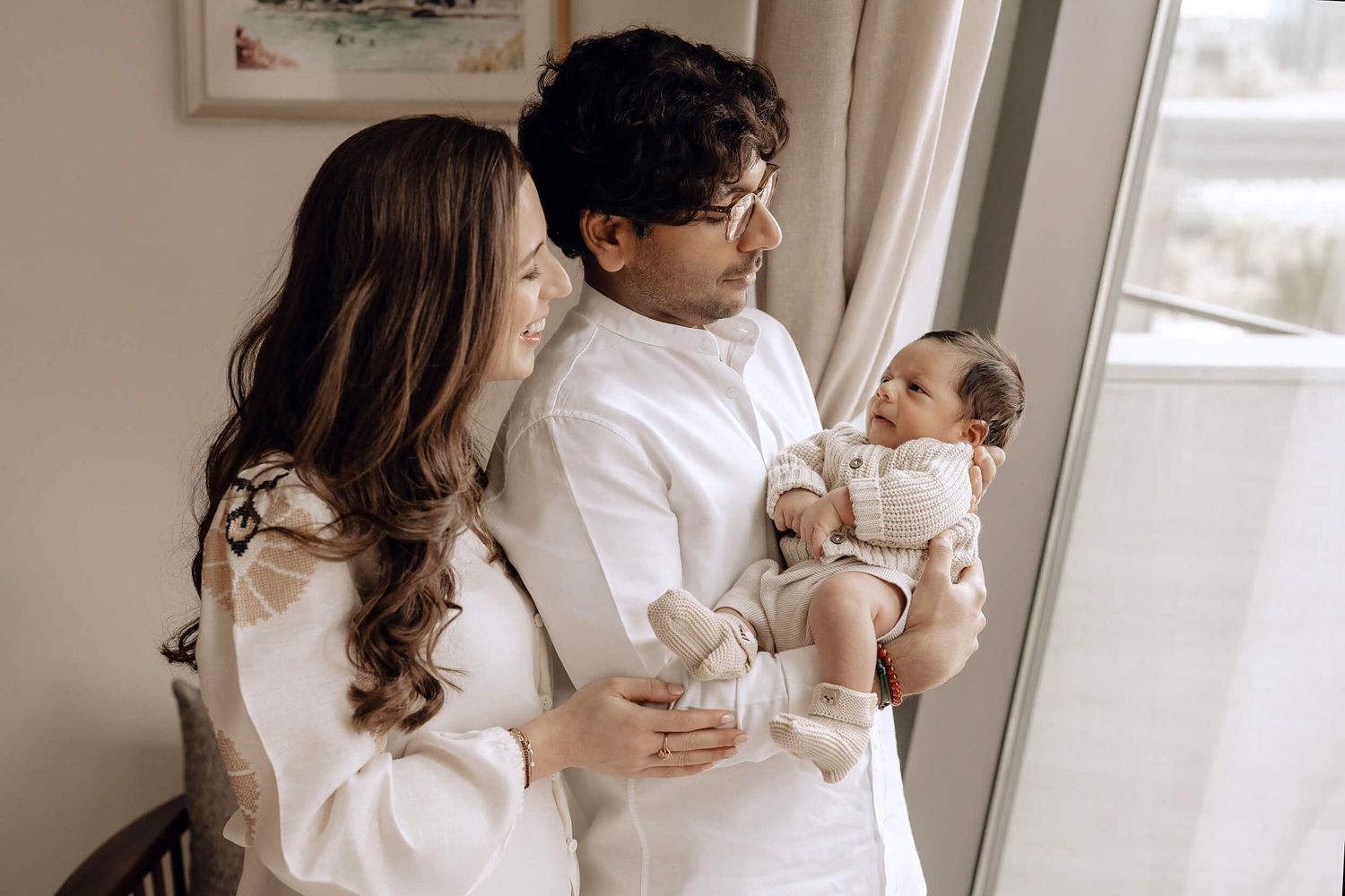 A couple in matching white outfits tenderly holding their baby near a bright window.