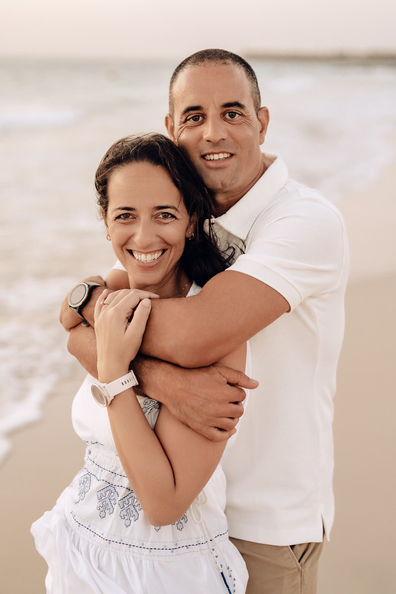 Romantic couple embracing on a beach at sunset wearing white and beige outfits.