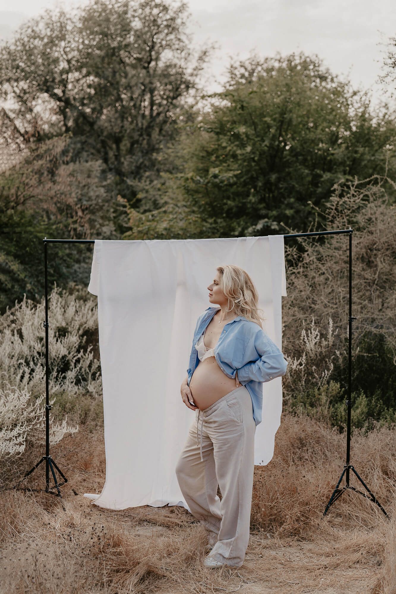 A maternity photo shoot outdoors with a white backdrop showing a pregnant figure in a blue shirt and beige pants.