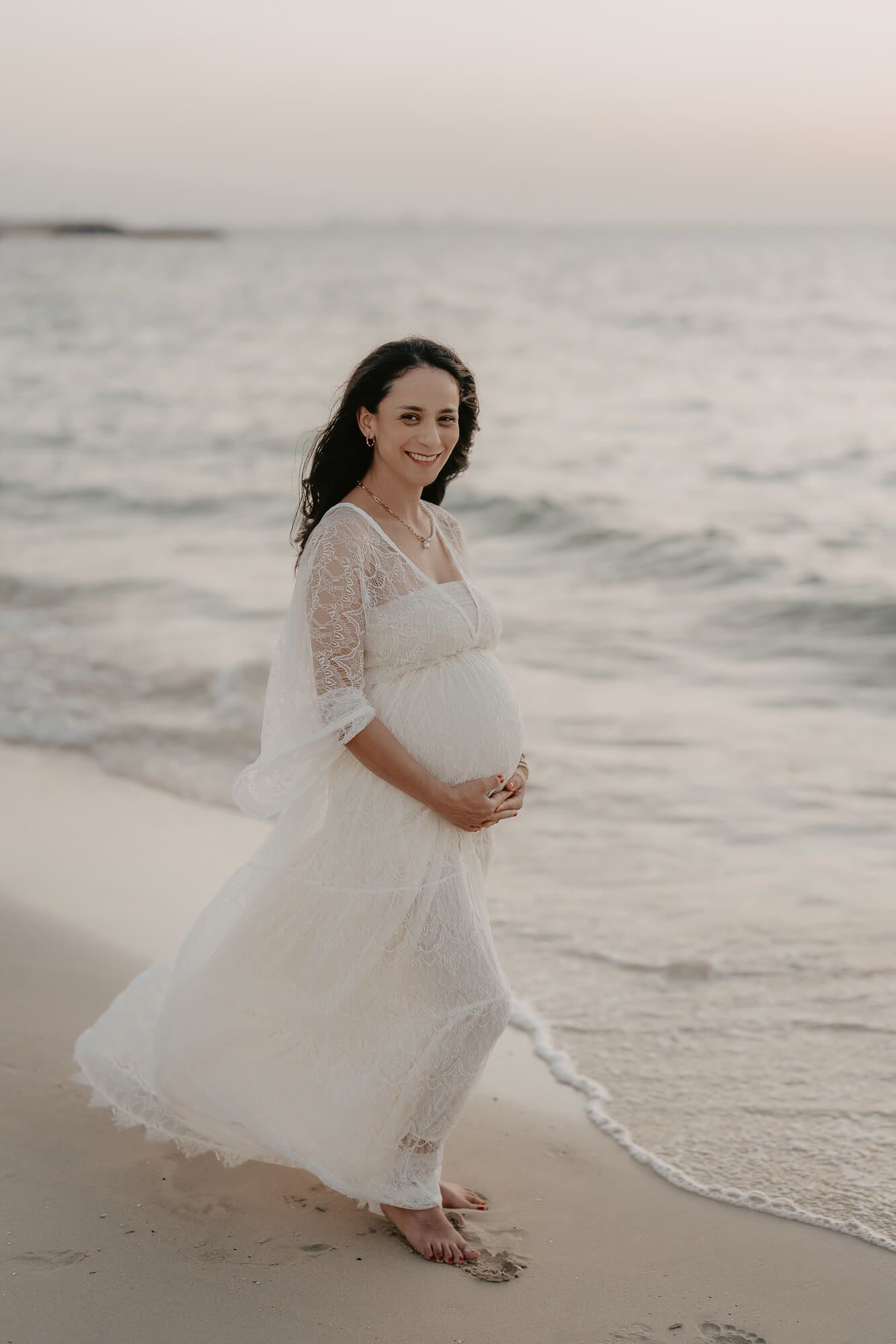 A pregnant woman in a flowing white lace dress poses gracefully on a serene beach at sunset, creating romantic coastal imagery.