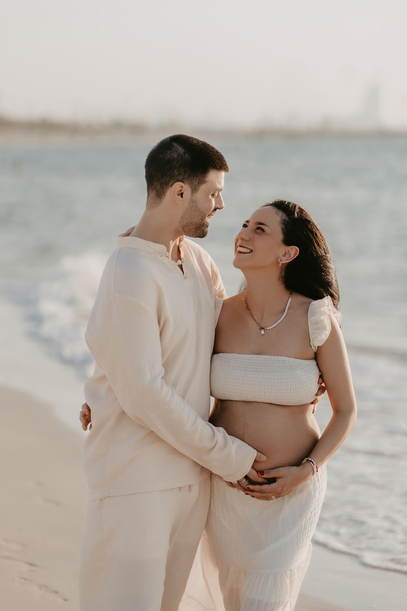 A couple embraces and laughs together on a sunny beach in white outfits during a romantic photoshoot session.