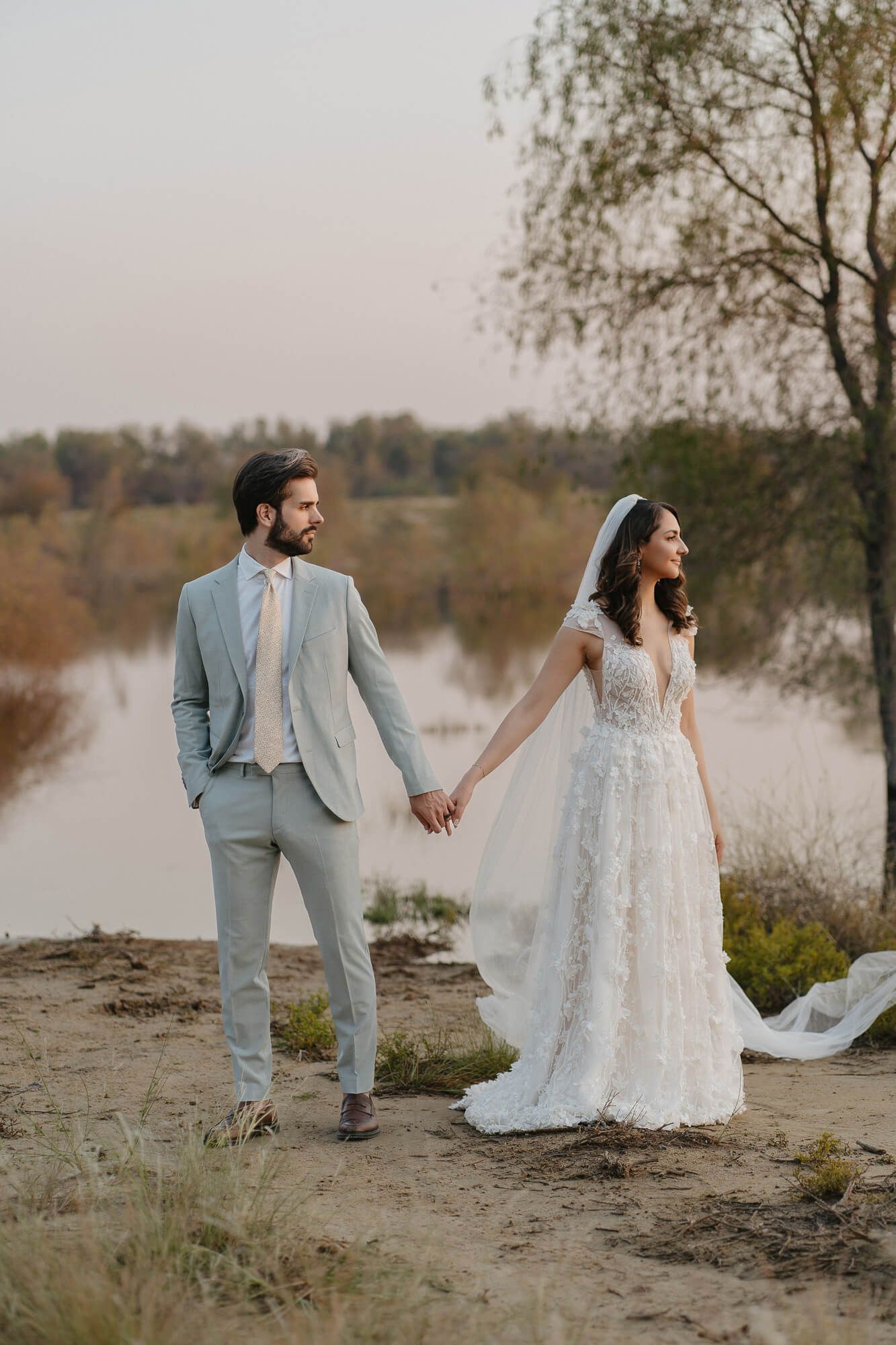 A couple holds hands during their lakeside wedding photo session in romantic muted tones at sunset.