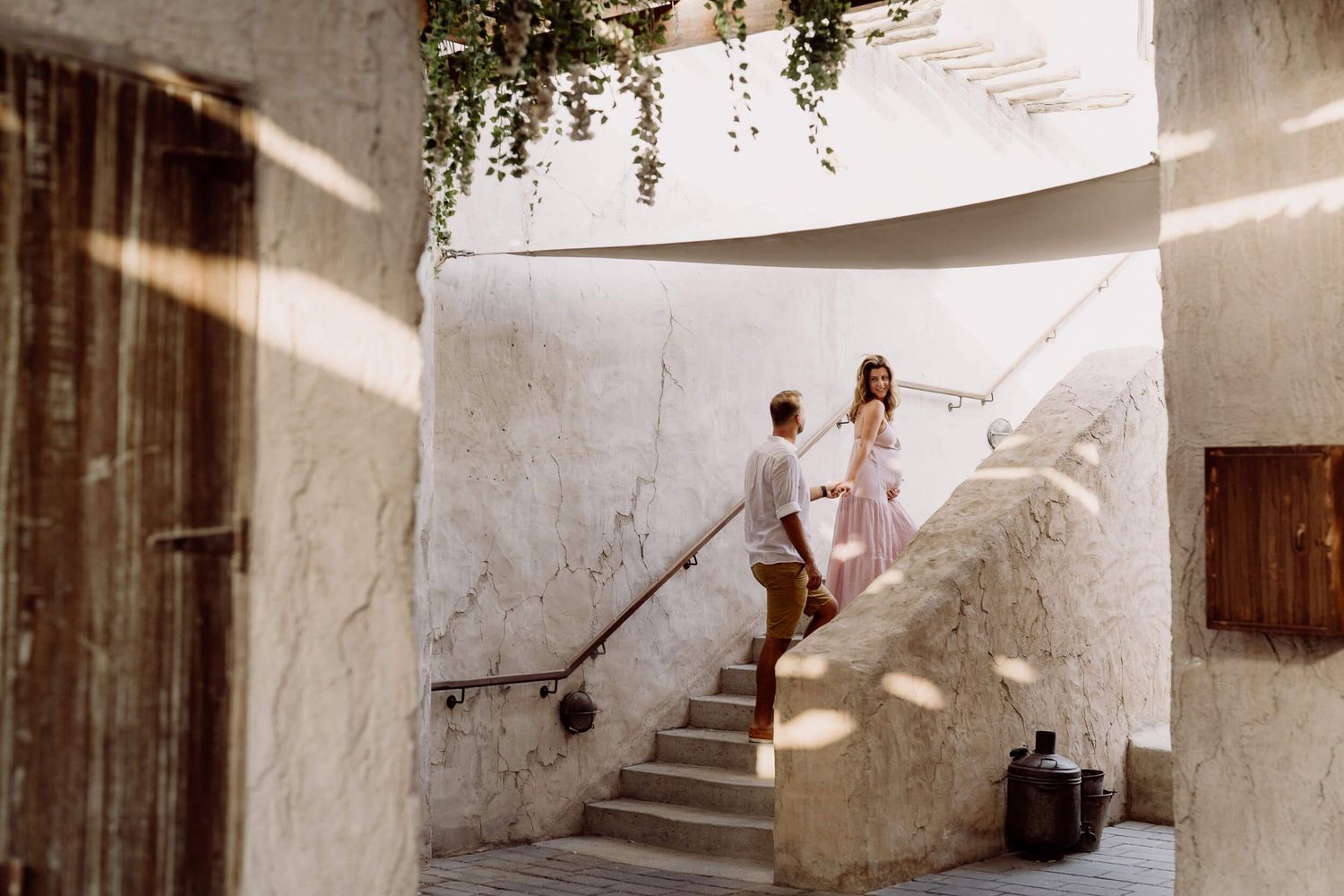 Two people share a romantic moment on white stone stairs in a Mediterranean courtyard with hanging plants.