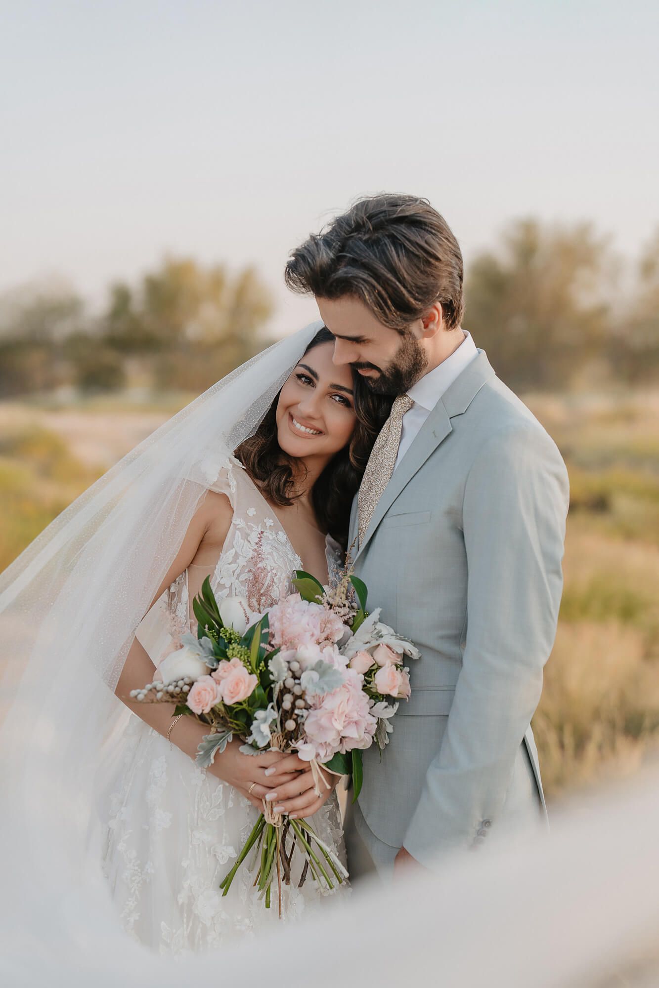 A romantic wedding moment captured in a field with a beautiful bouquet of pink and white flowers and a light gray suit.