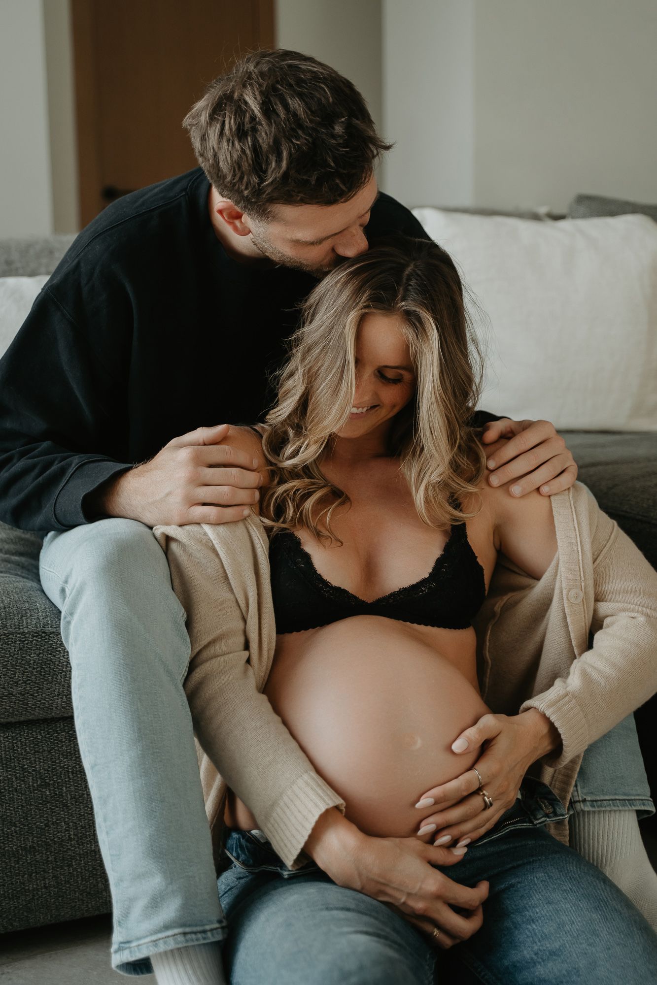 Intimate maternity photo of couple sitting on couch showing pregnant belly in black bra and beige cardigan.