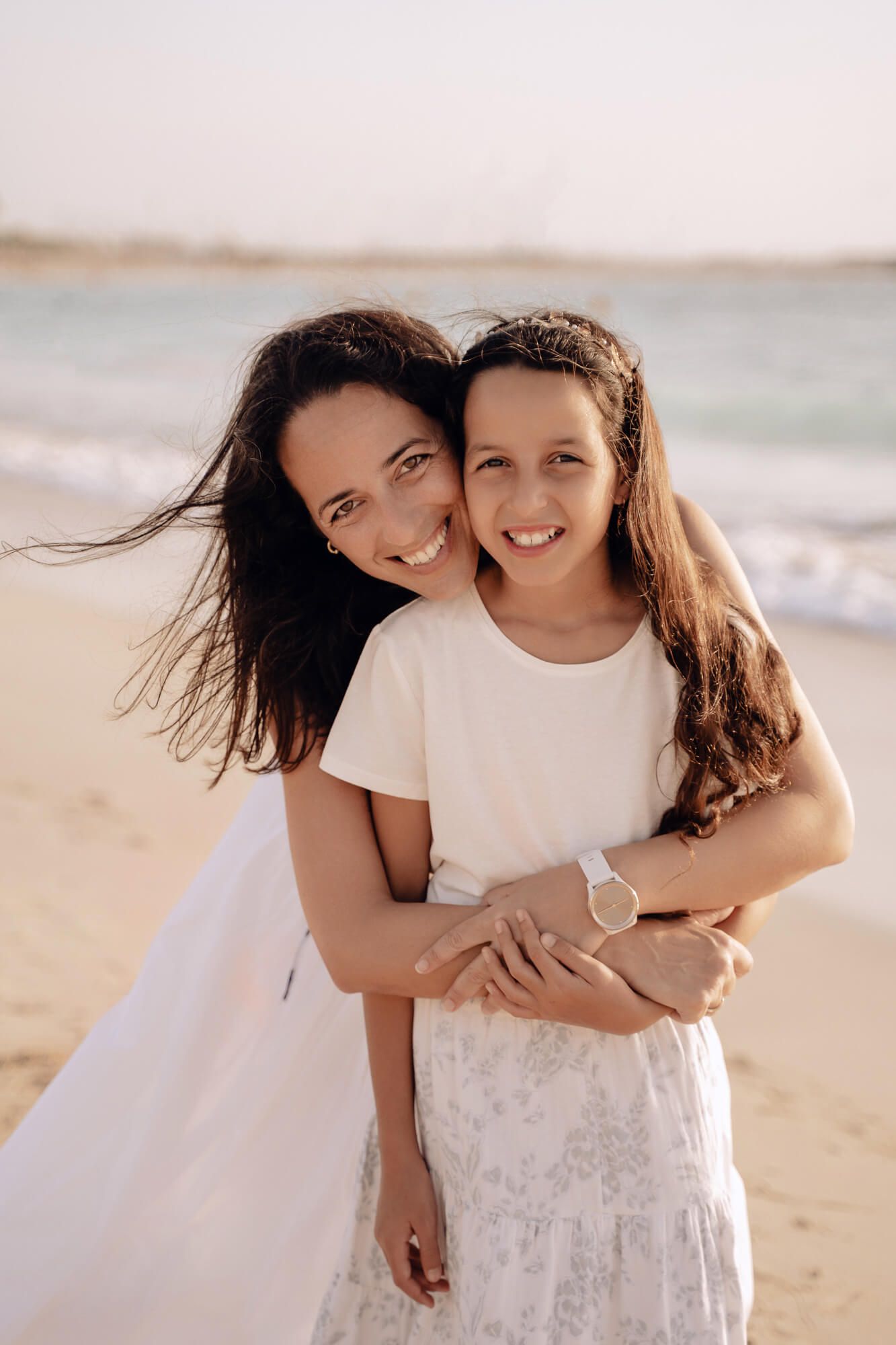 Two people share a warm embrace while wearing white outfits on a sunny beach.