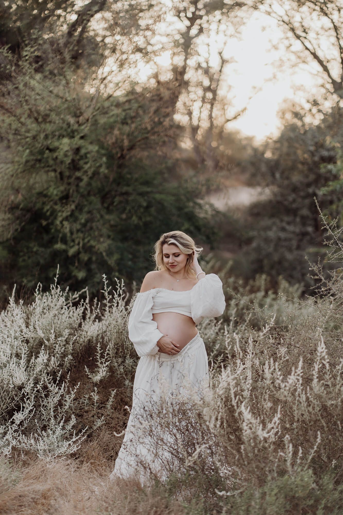 A pregnant woman in an off-shoulder white dress poses in a field of sage brush.