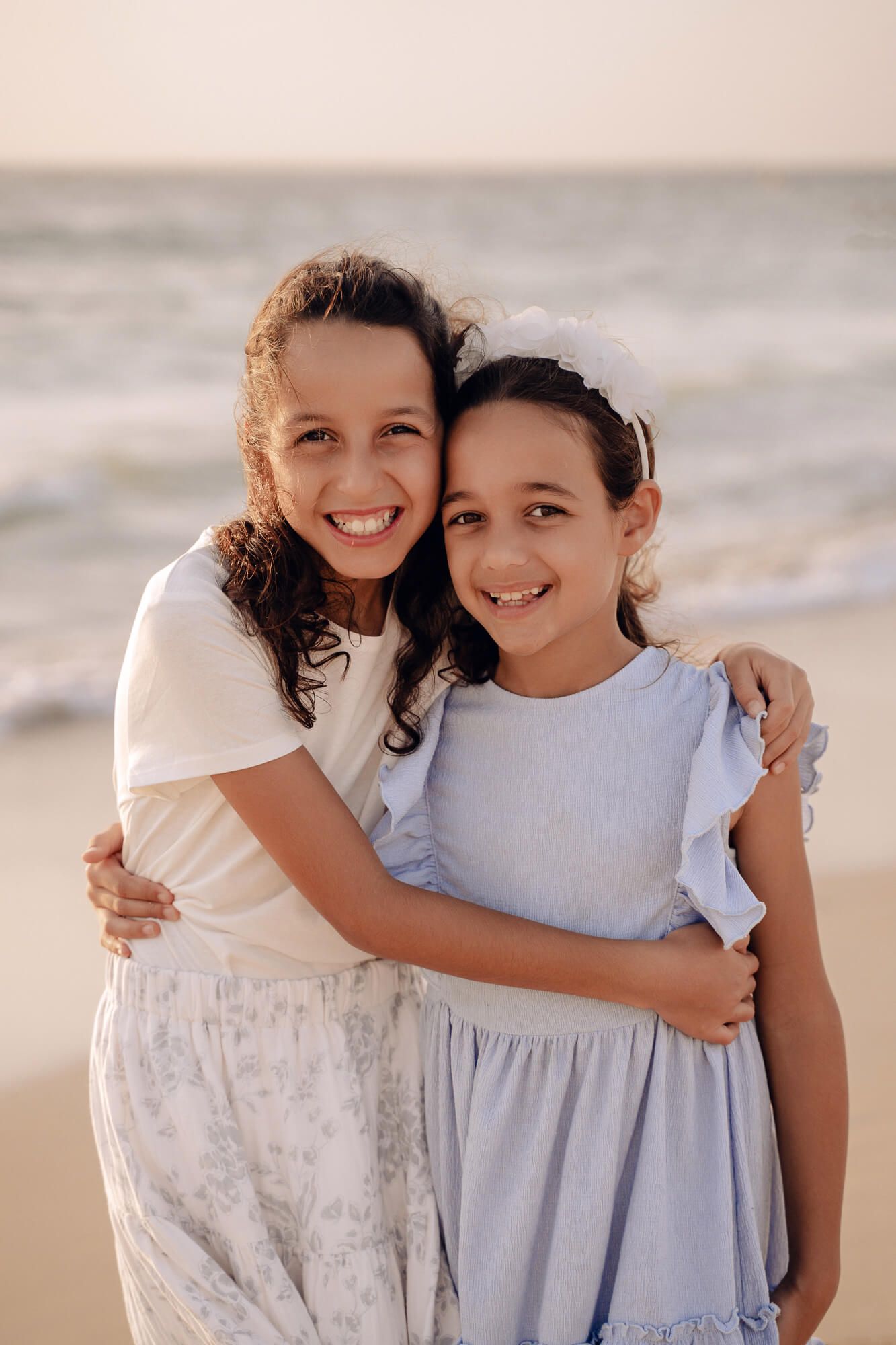Two children in white and blue dresses hugging and smiling on the beach.