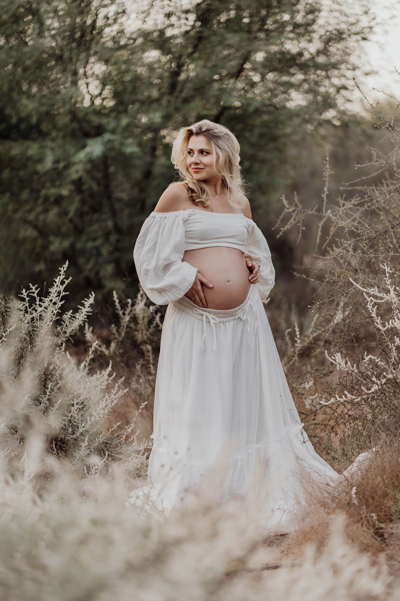 Dreamy maternity session in white dress among wild grass and trees.