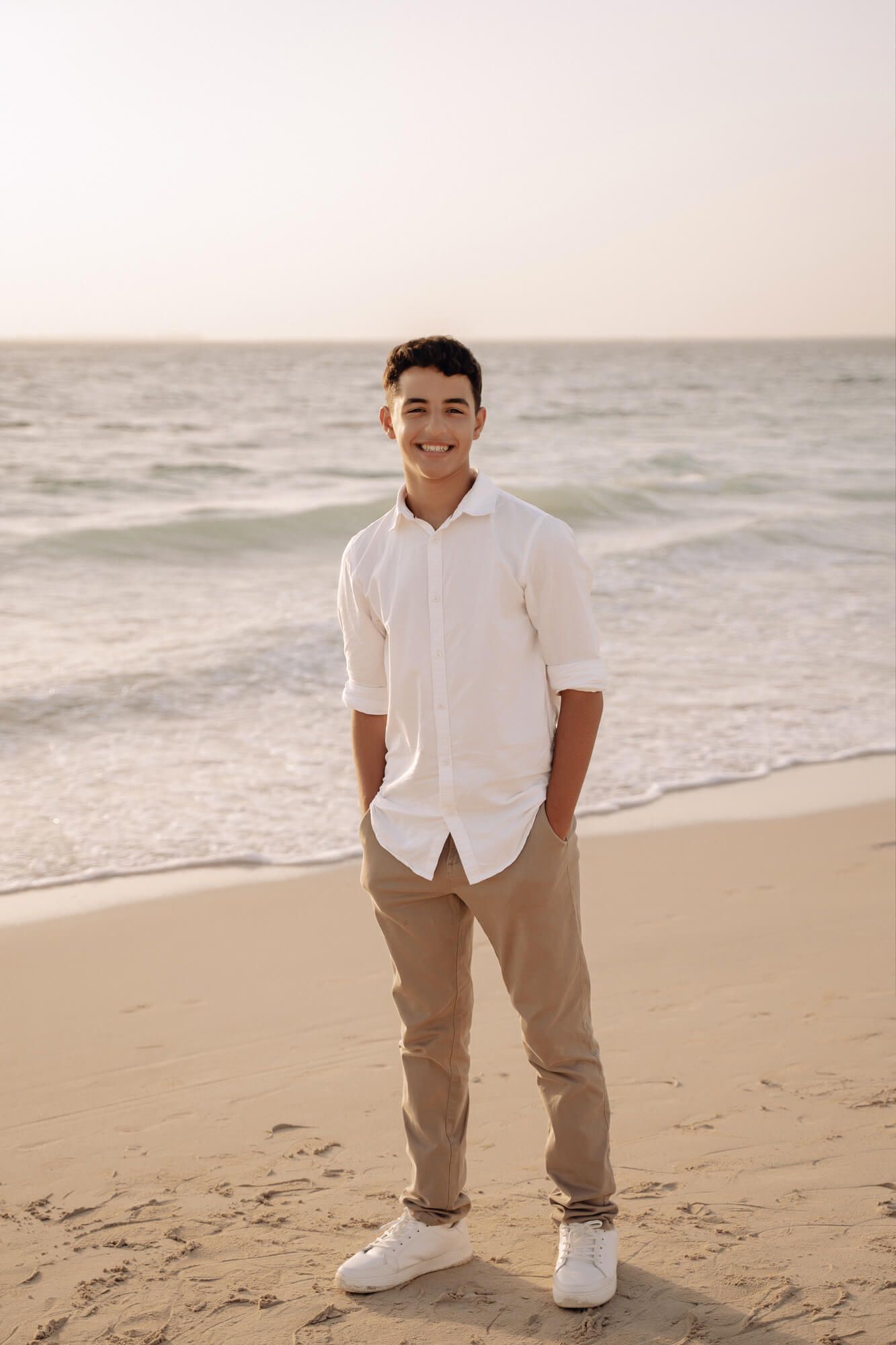 Person in casual white shirt and khakis poses on sandy beach at golden hour.