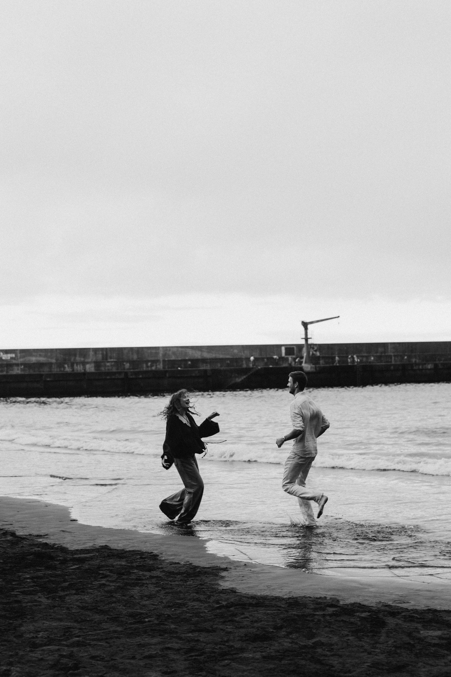 Two people play and run joyfully along a beach near a seawall in black and white photography.