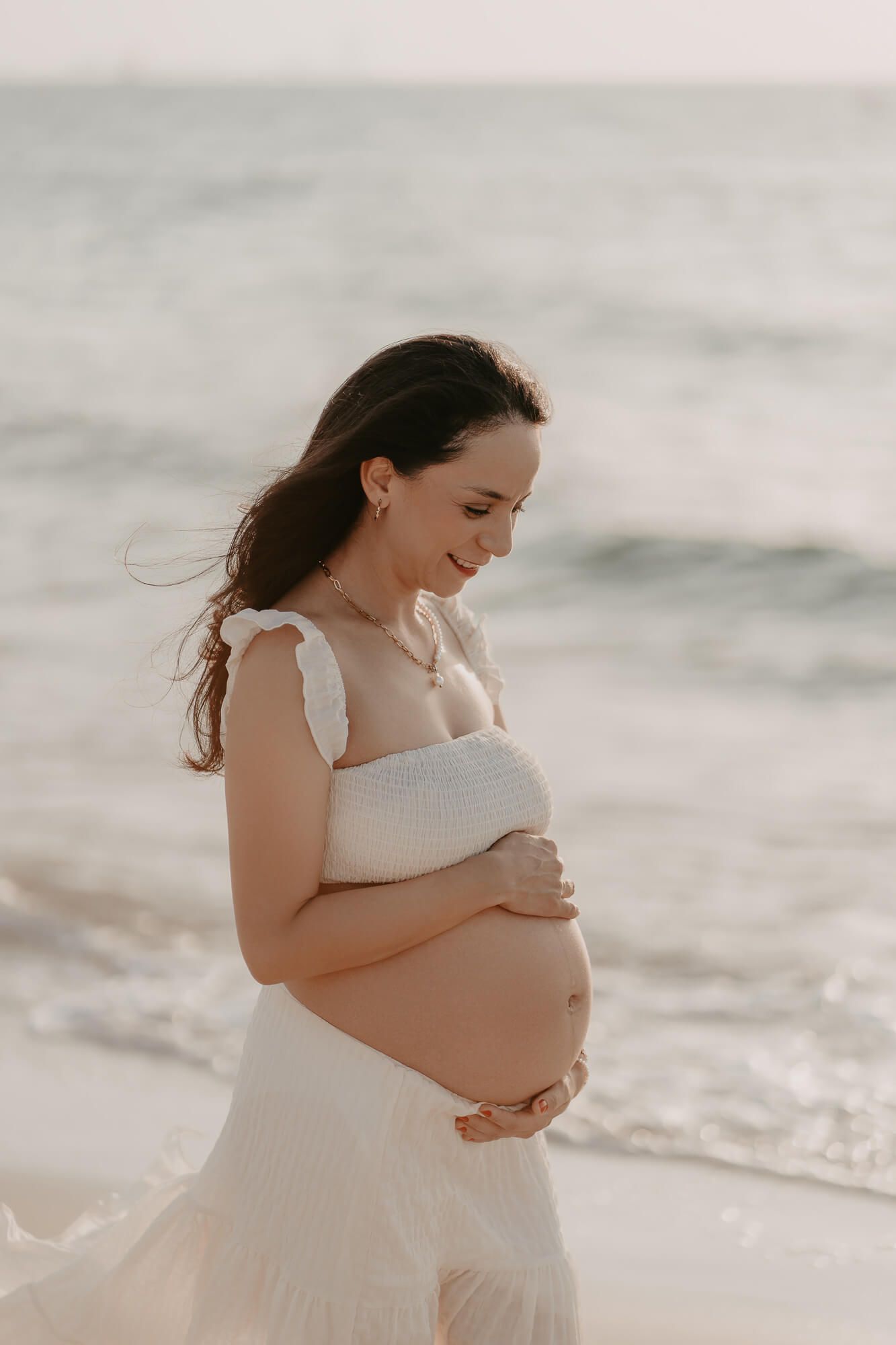 A pregnant woman in a white dress stands on a serene beach at sunset, gently cradling her belly.