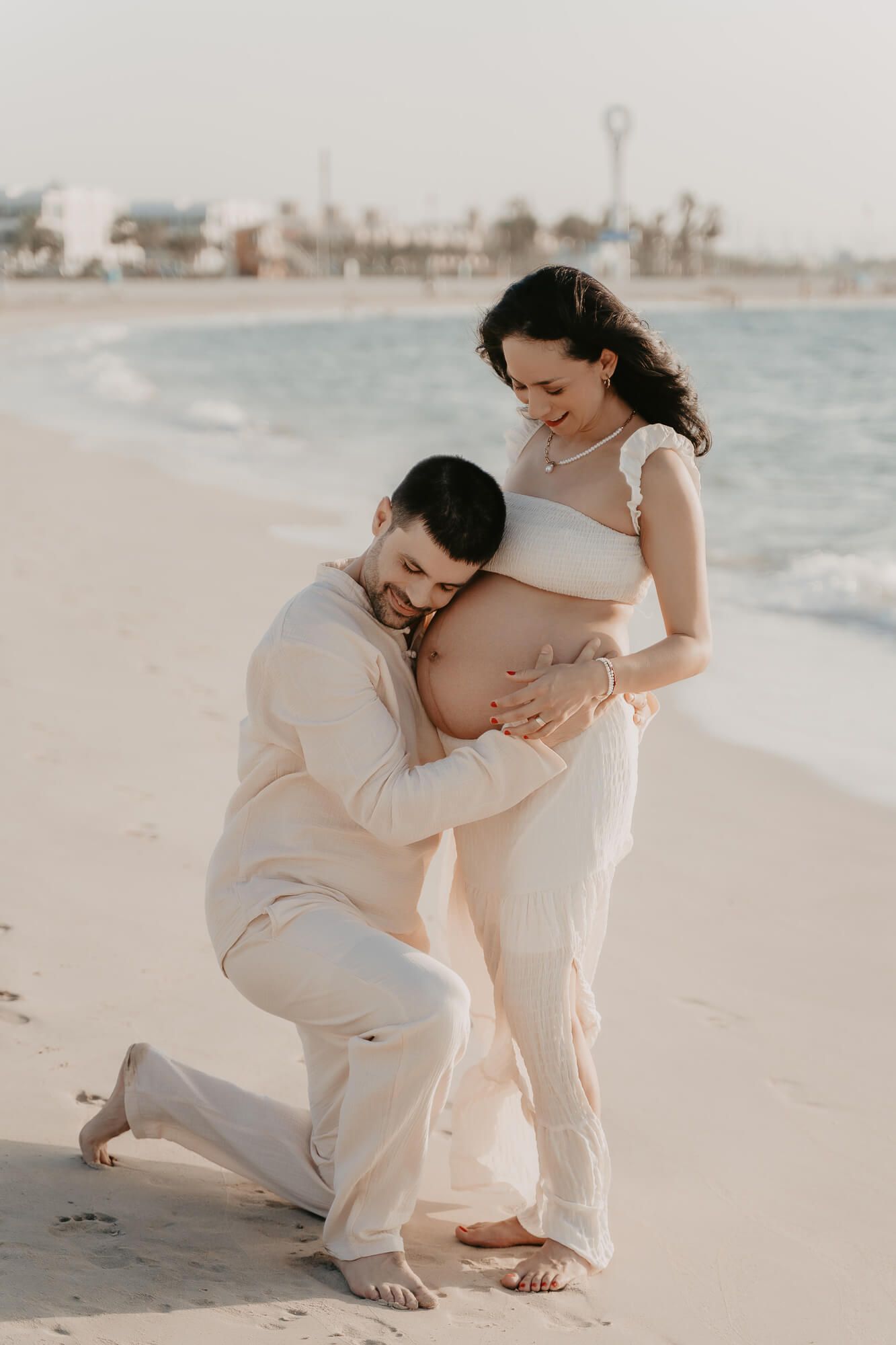 A tender maternity moment captured on a beach at sunset, with a couple in light colored clothing embracing on the sand.