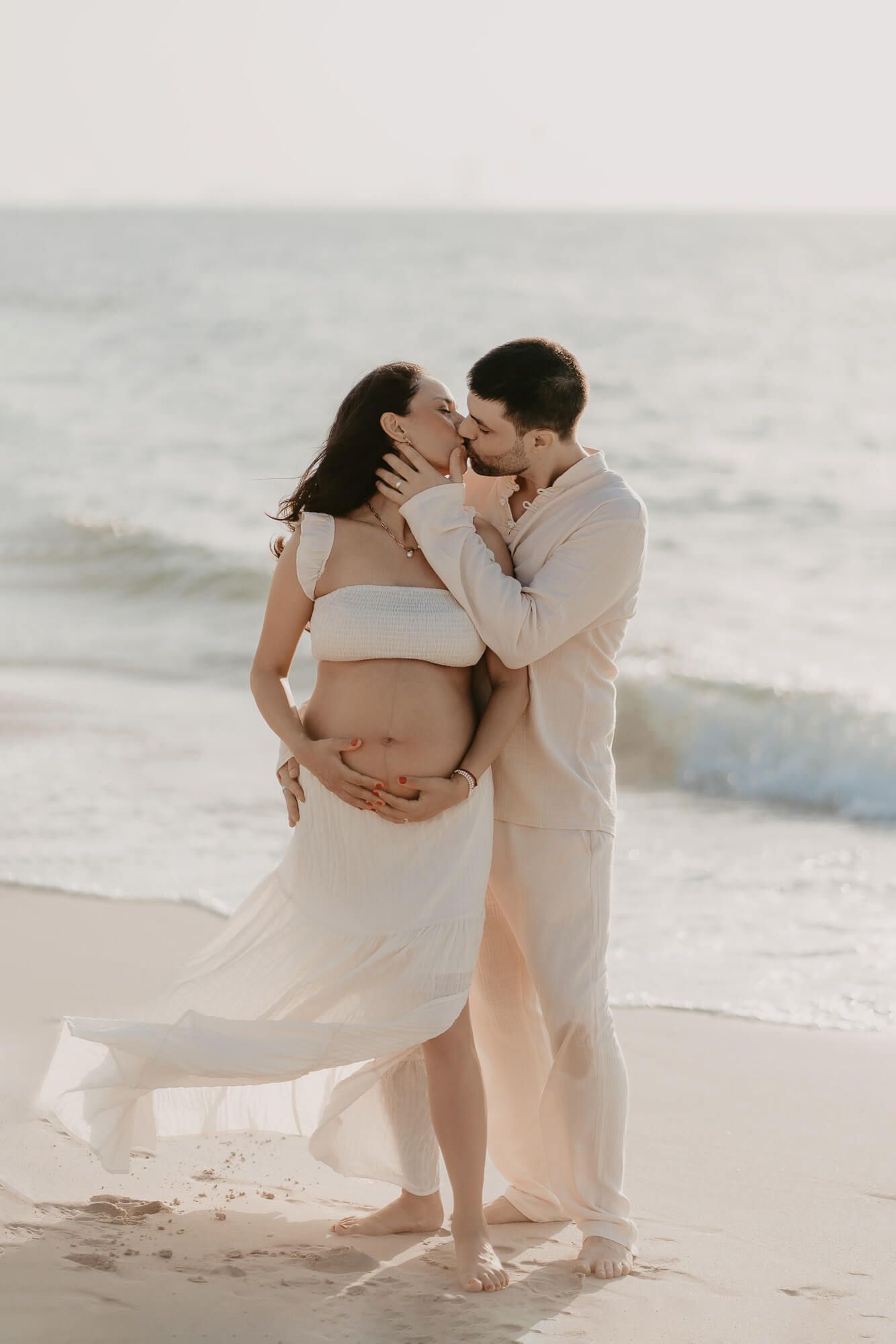 Romantic maternity beach photoshoot with flowing white dress and soft ocean waves in the background.