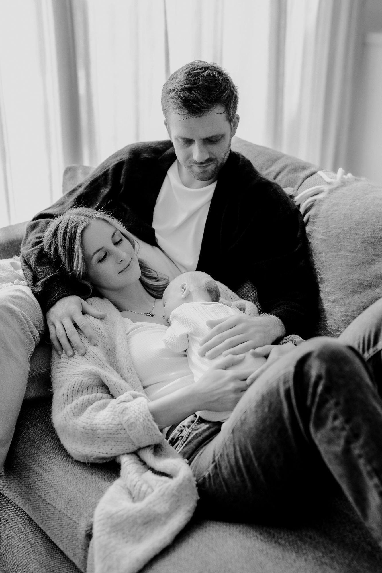 Black and white intimate moment of parents cuddling with their newborn baby on a cozy couch.