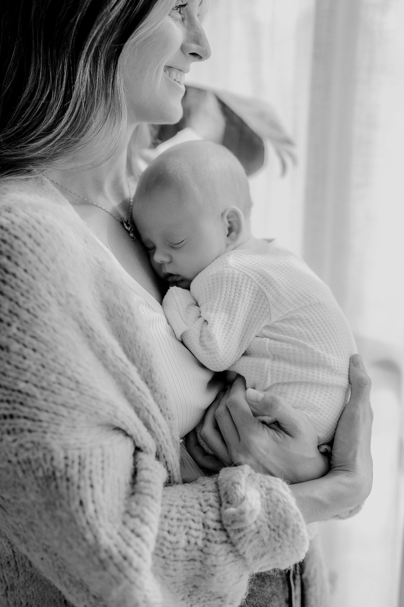 Black and white intimate portrait of a tender moment between parent and sleeping baby in cozy sweaters by window light.