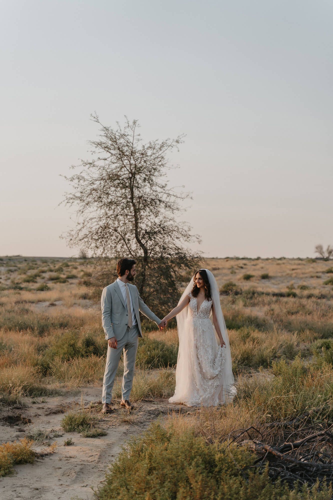 A couple in wedding attire stands together in a desert landscape at dusk with a lone tree in the background.