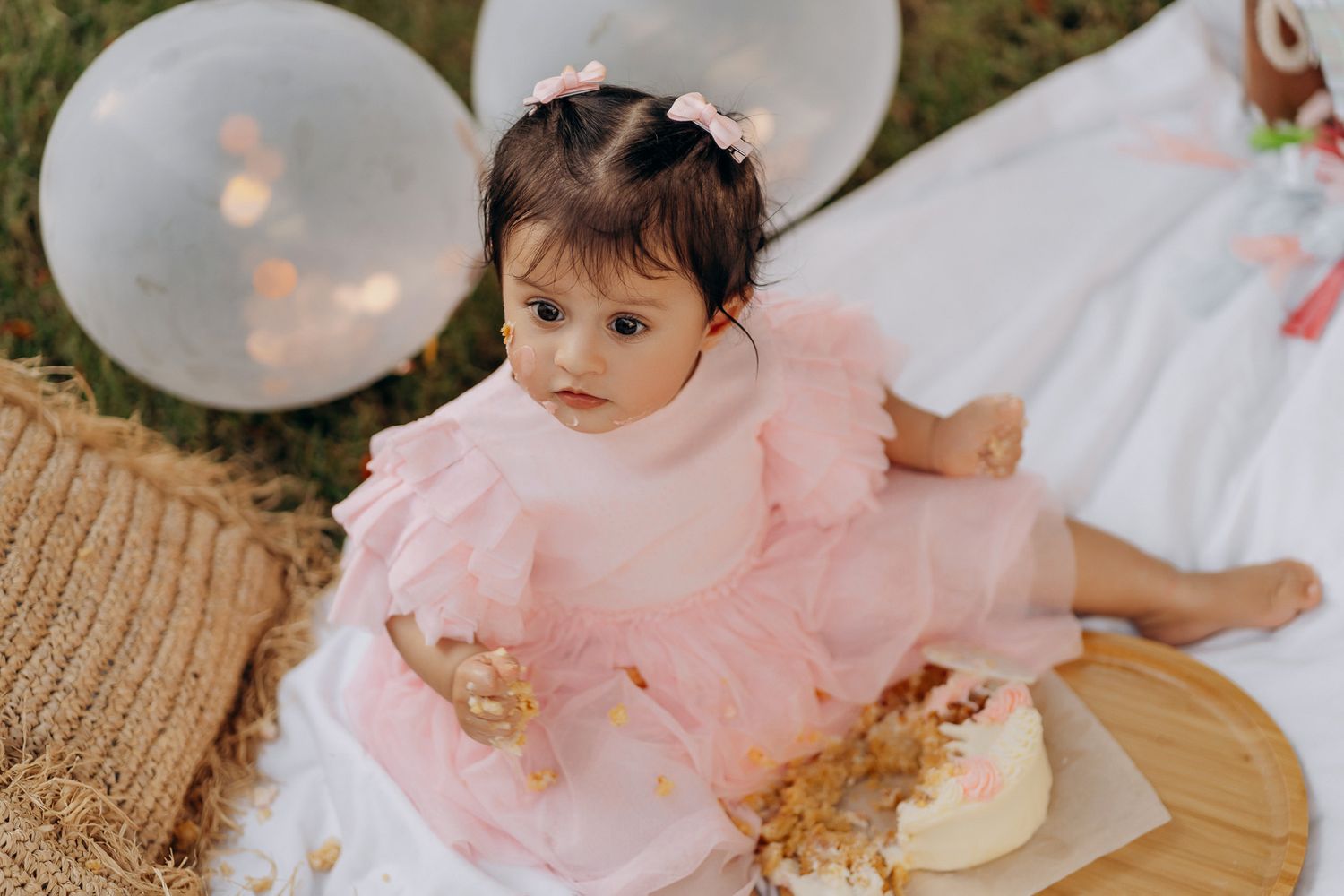 Baby wearing a pink tulle dress sits on blanket with white balloons at outdoor celebration.