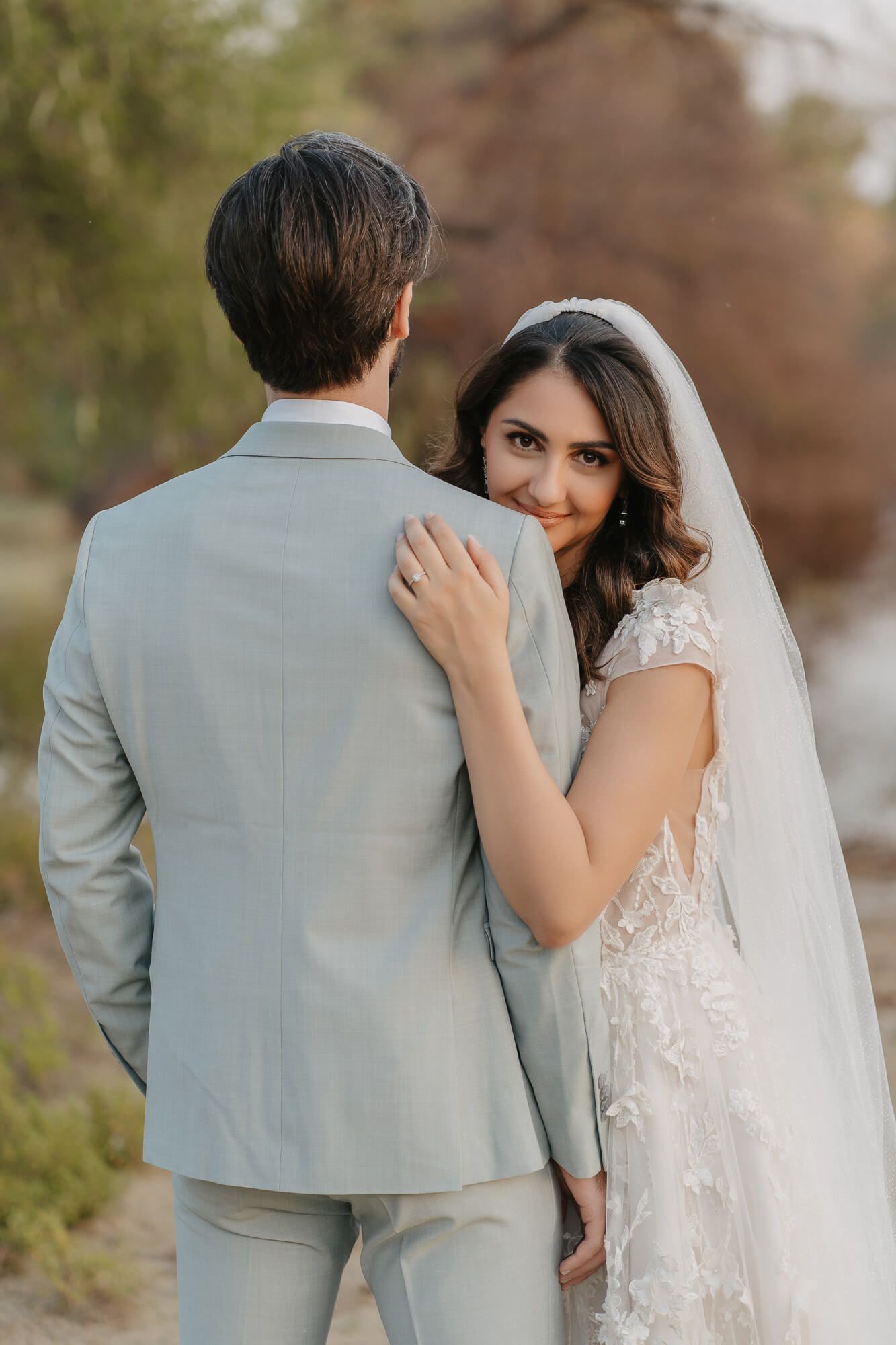 A tender moment between newlyweds with the bride in lace and veil embracing her groom in a light gray suit.