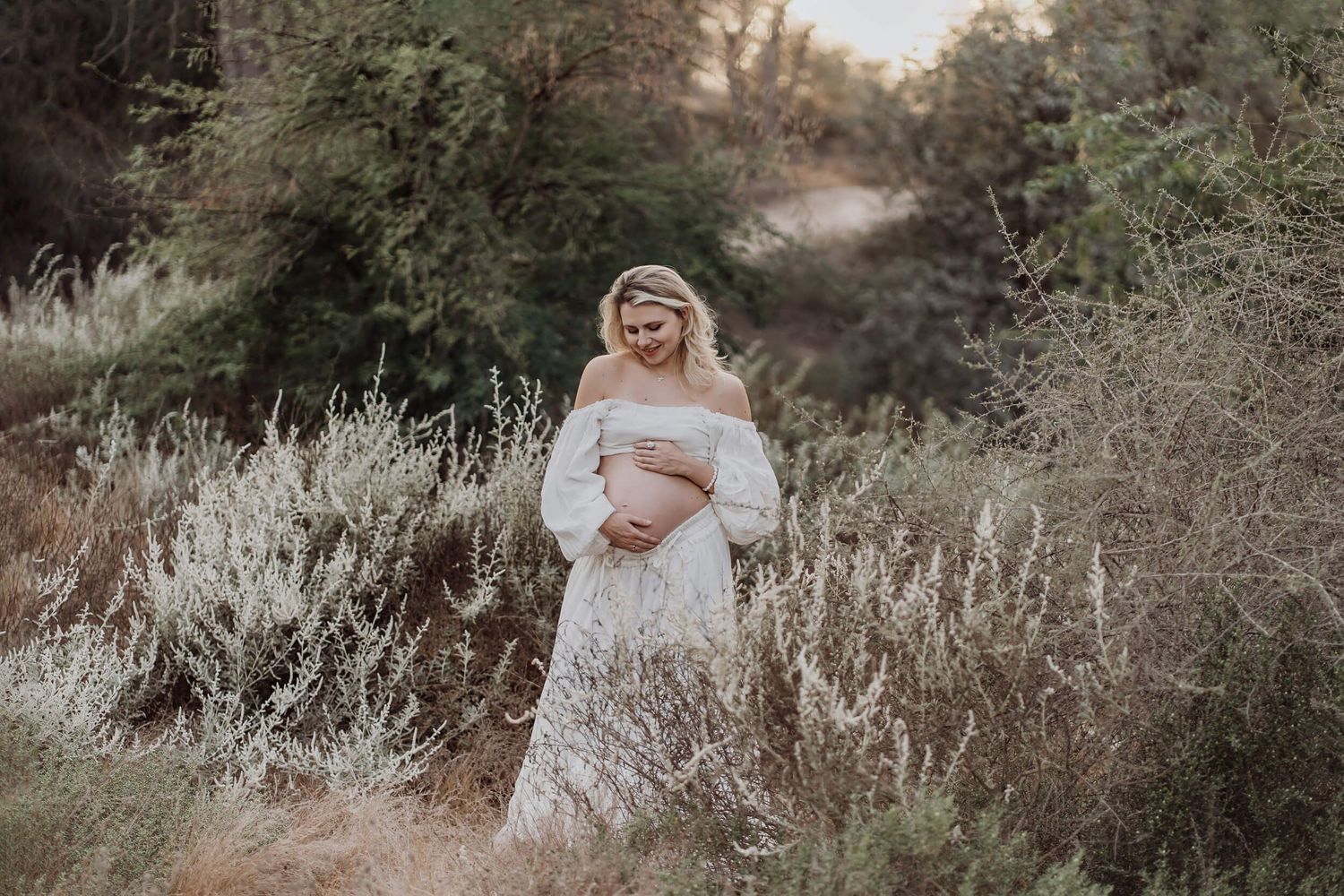 A joyful maternity photo of a woman in white dress surrounded by natural foliage.