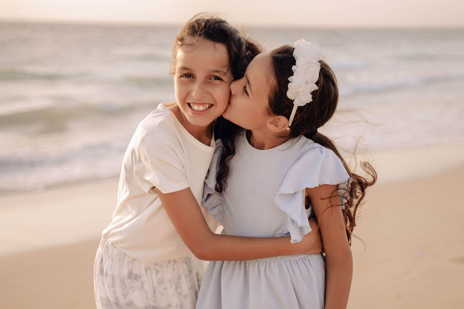 Two children embracing in light colored dresses during sunset beach photography.